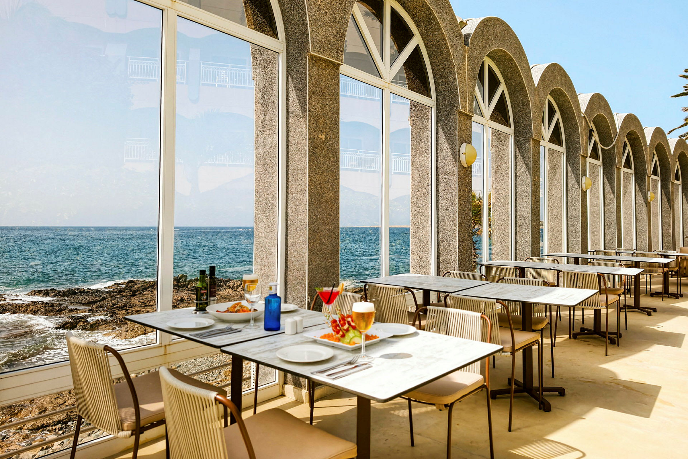 Restauranger med bord uppställt bredvid stora glasfönster som vetter mot havet. (AI-genererad text)