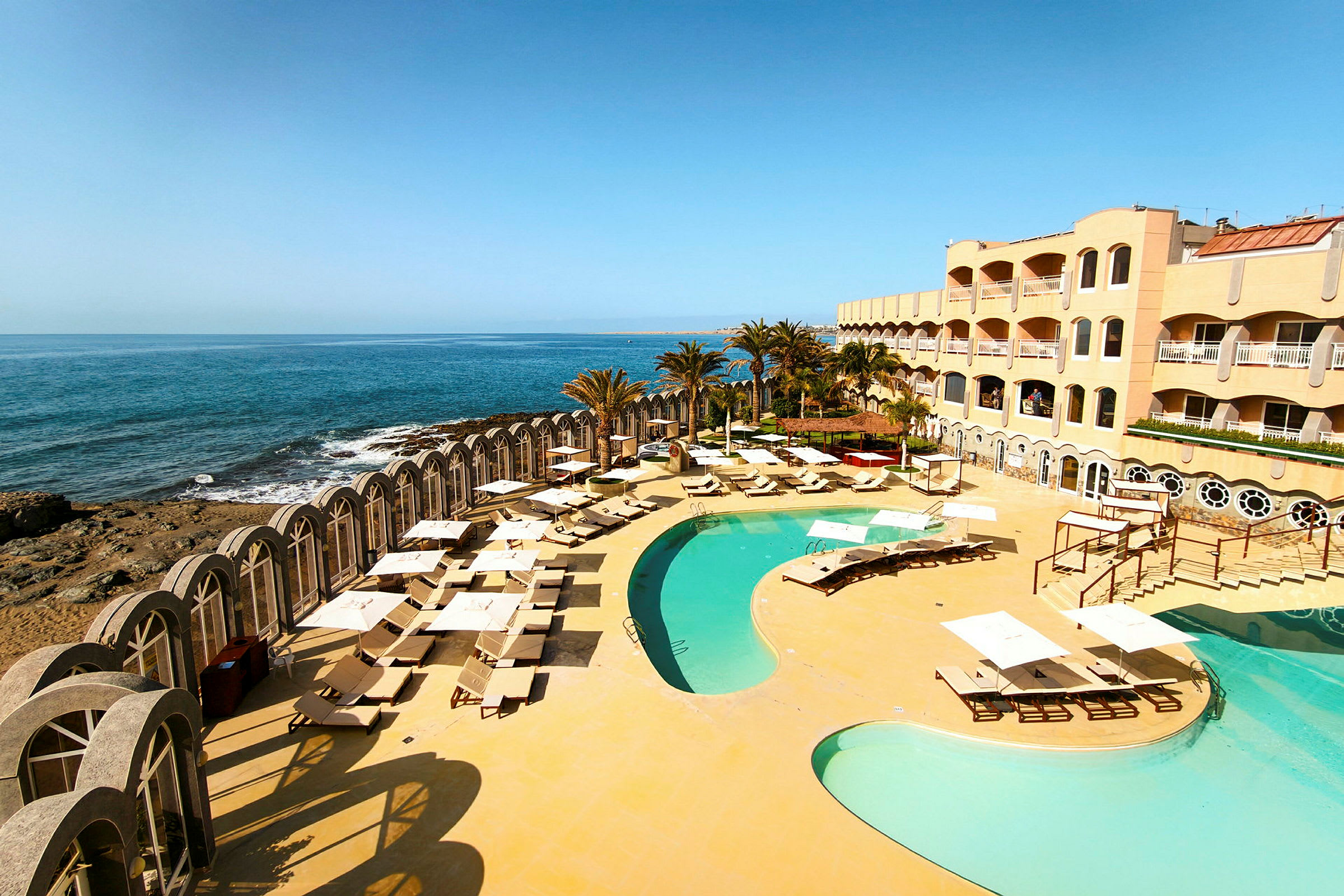 Bild av ett hotellkomplex med pool och solstolar vid havet. (AI-genererad text)
