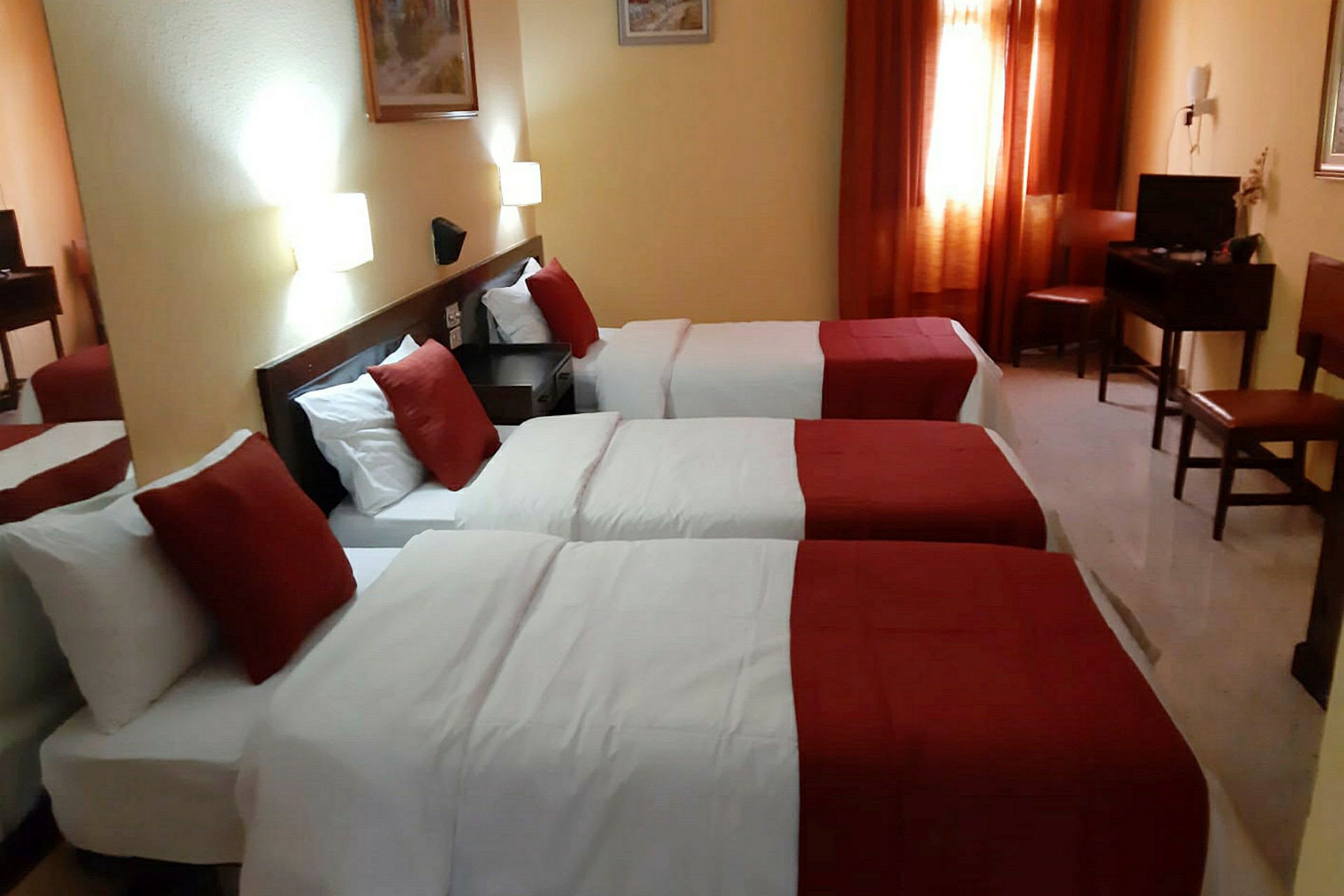 Ett hotellrum med tre enkelsängar, vita lakan och röda överkast, med ett nattduksbord och belysning på väggen. (AI-genererad text)