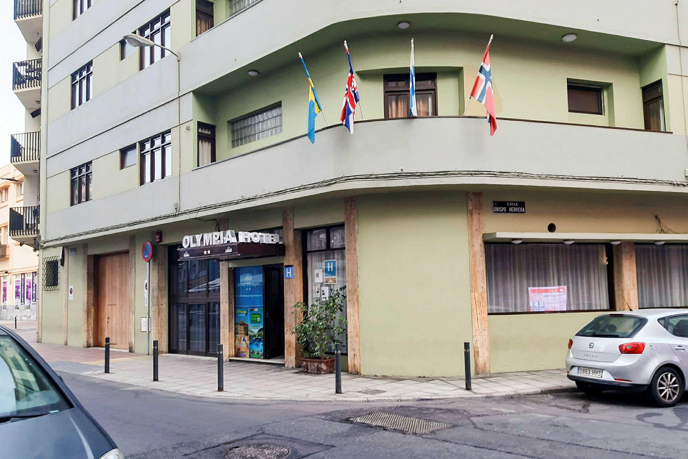 En grön byggnad med texten 'Olympia Hotel' ovanför ingången. Flera flaggor är uppsatta på en balkong. (AI-genererad text)