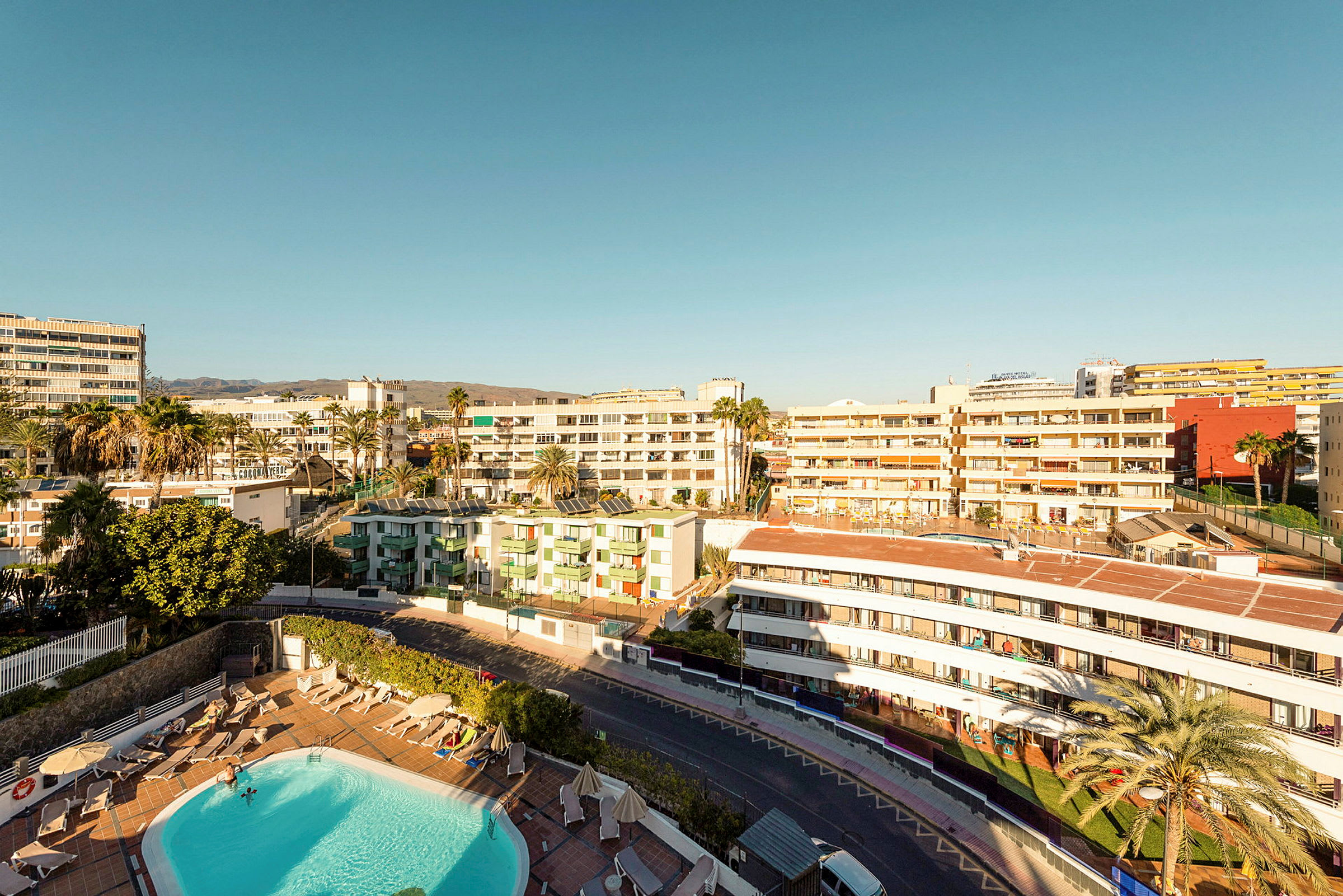 Vy över ett hotellområde med pool, omgiven av byggnader under en klar himmel. (AI-genererad text)