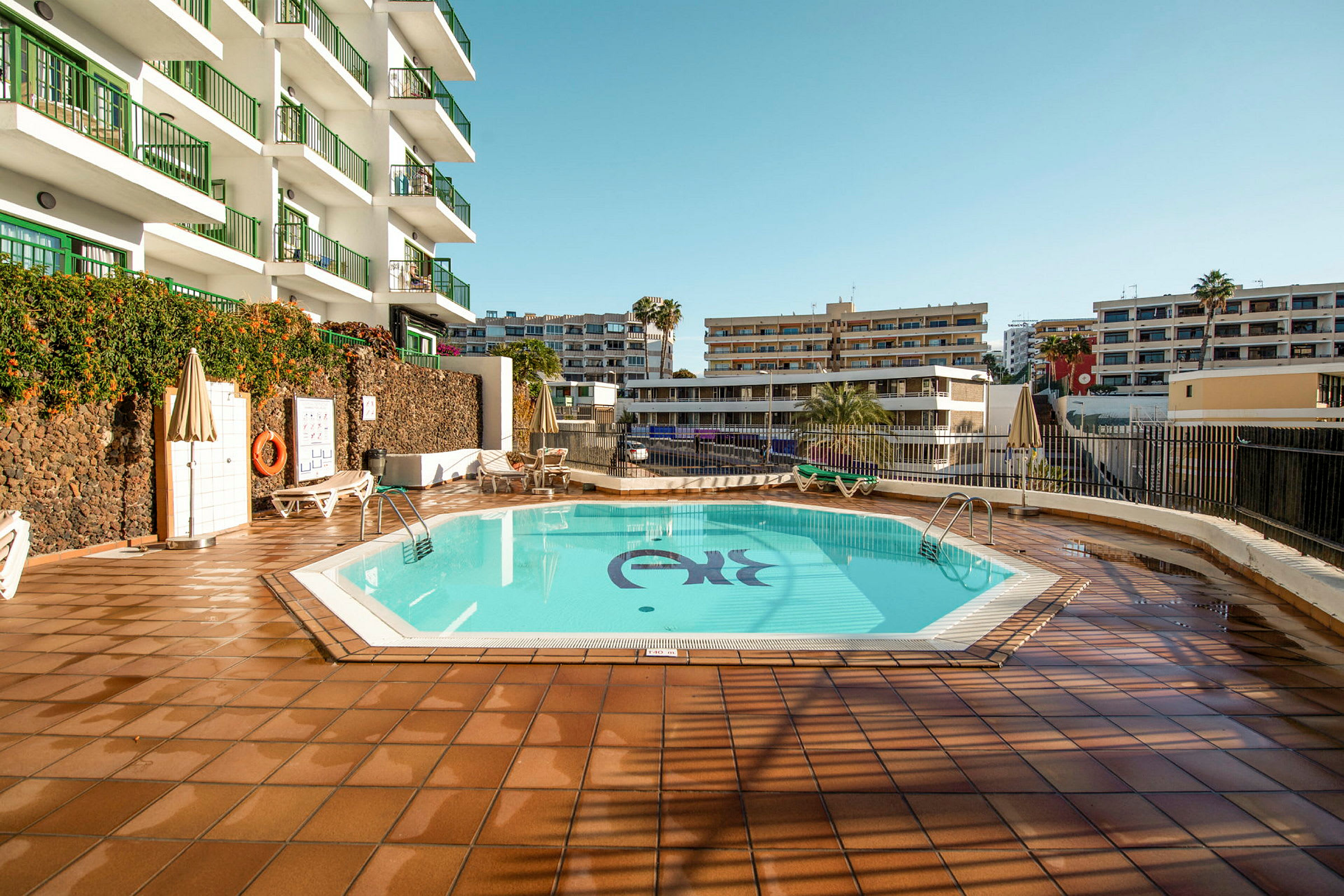 En hotellpool omgiven av solstolar och byggnader. (AI-genererad text)