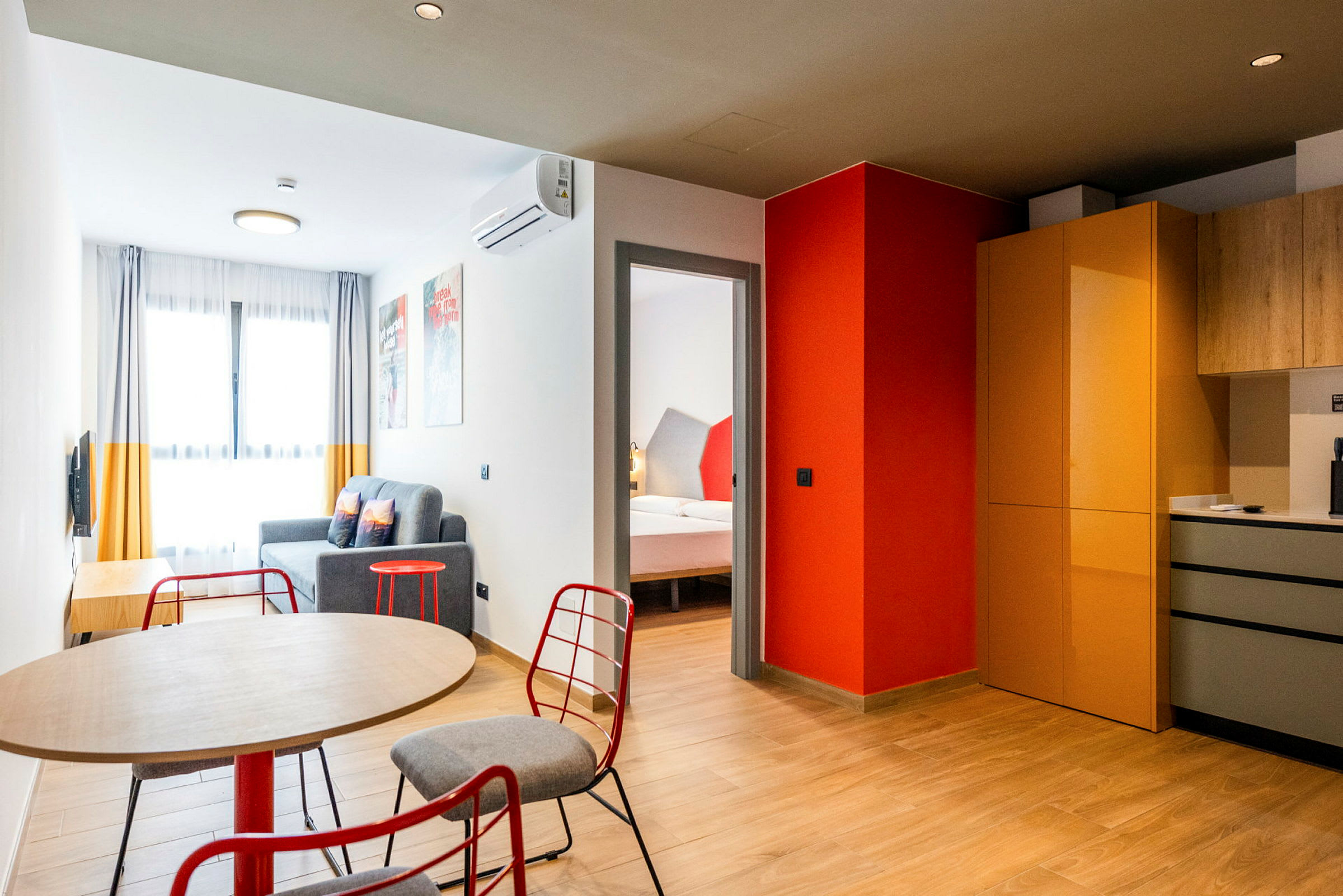 Ett modernt inrett hotellrum med ett bord och stolar i förgrunden, en sittgrupp med soffa och ett sovrum synligt genom en dörröppning. Köksdelen har skåp och arbetsytor. (AI-genererad text)