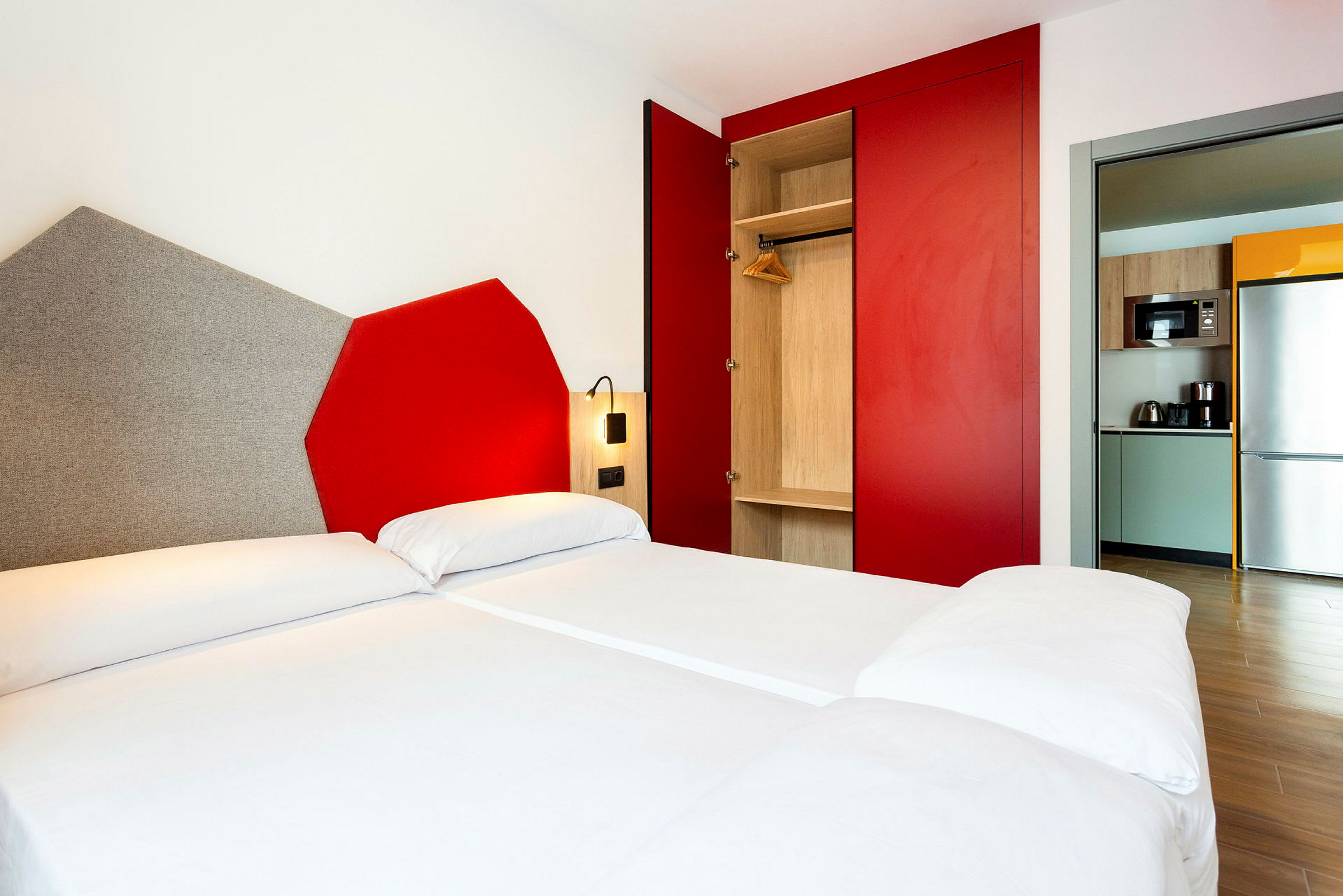 Bild av ett modernt hotellrum med en dubbelsäng, en garderob med röda skjutdörrar och en öppen liten köksyta med kylskåp och mikrovågsugn. (AI-genererad text)