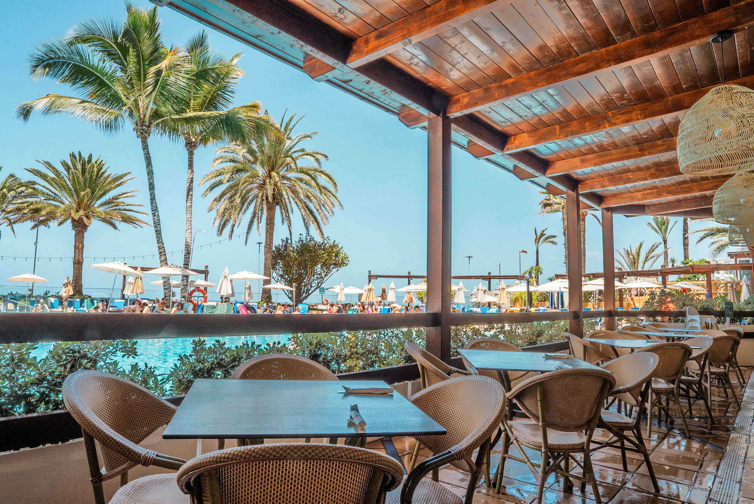 Utsikt från en träpergola med sittplatser mot en pool och palmer i bakgrunden. (AI-genererad text)