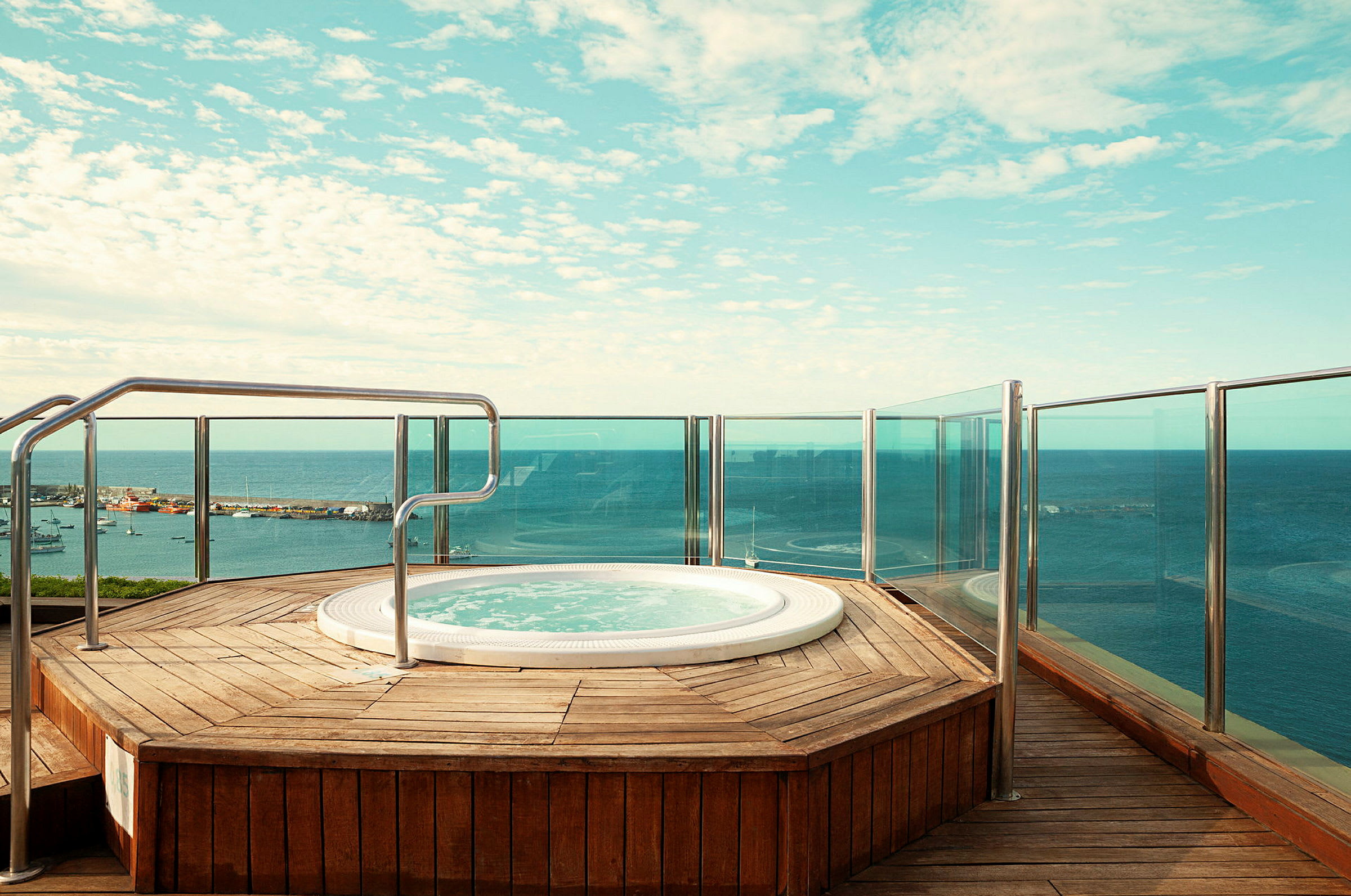 Bubbelpool på en träplattform med glasräcke och utsikt över havet. (AI-genererad text)
