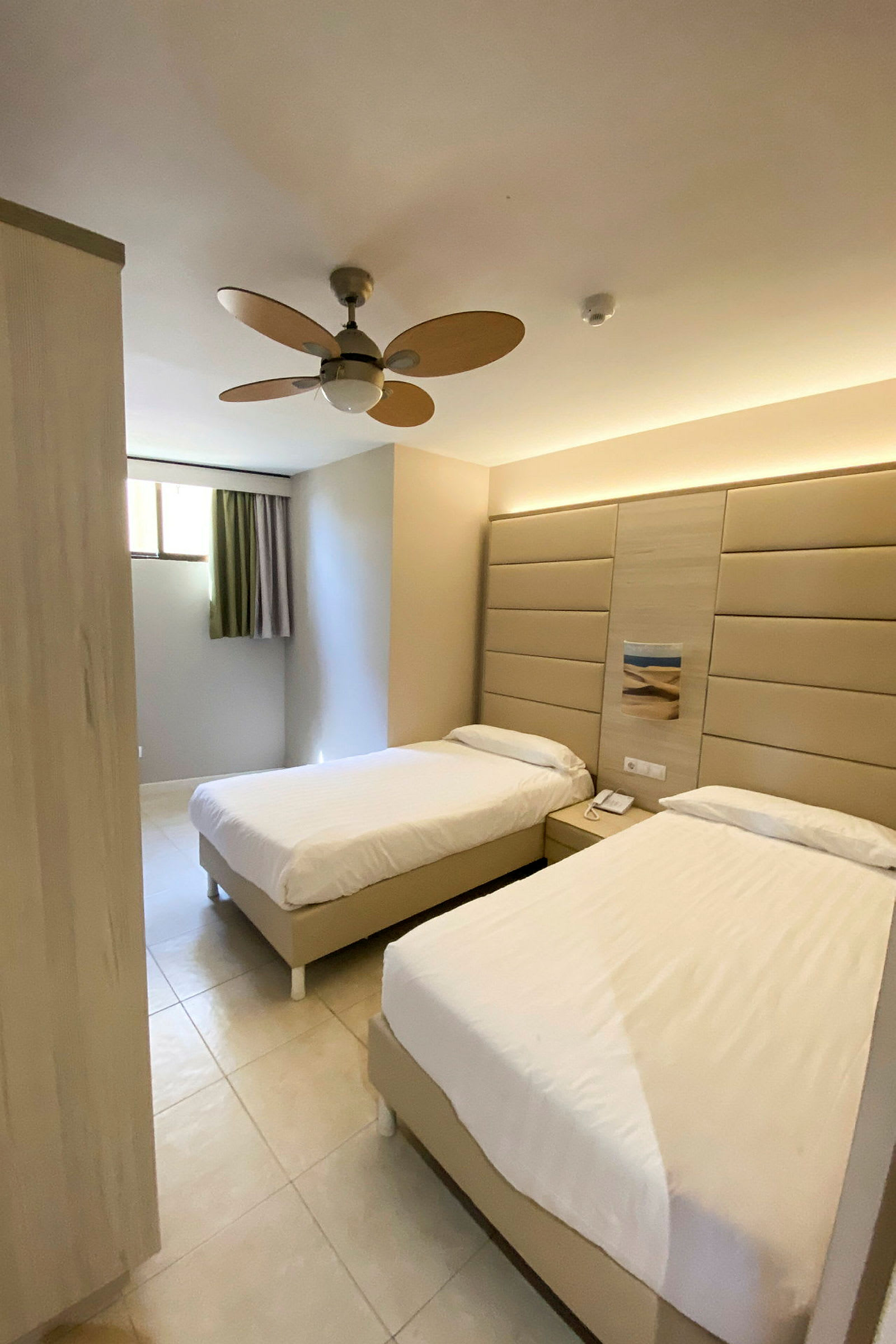Ett hotellrum med två enkelsängar, väggpanel bakom sängarna och ett takfläkt. (AI-genererad text)