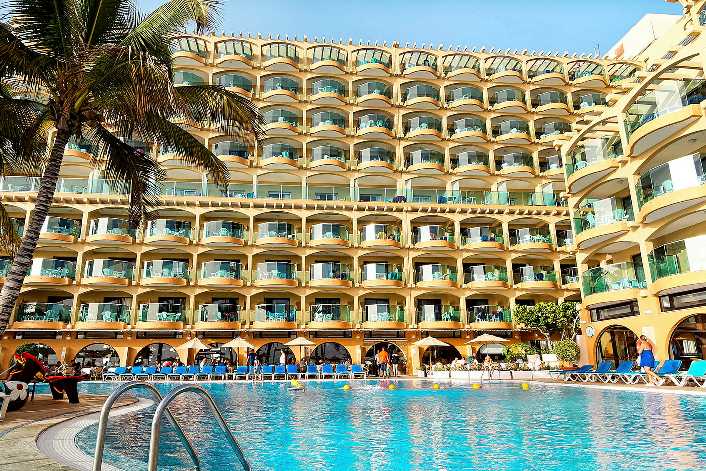 Hotellkomplex med flera våningar och balkonger, framför en pool med solsängar. (AI-genererad text)