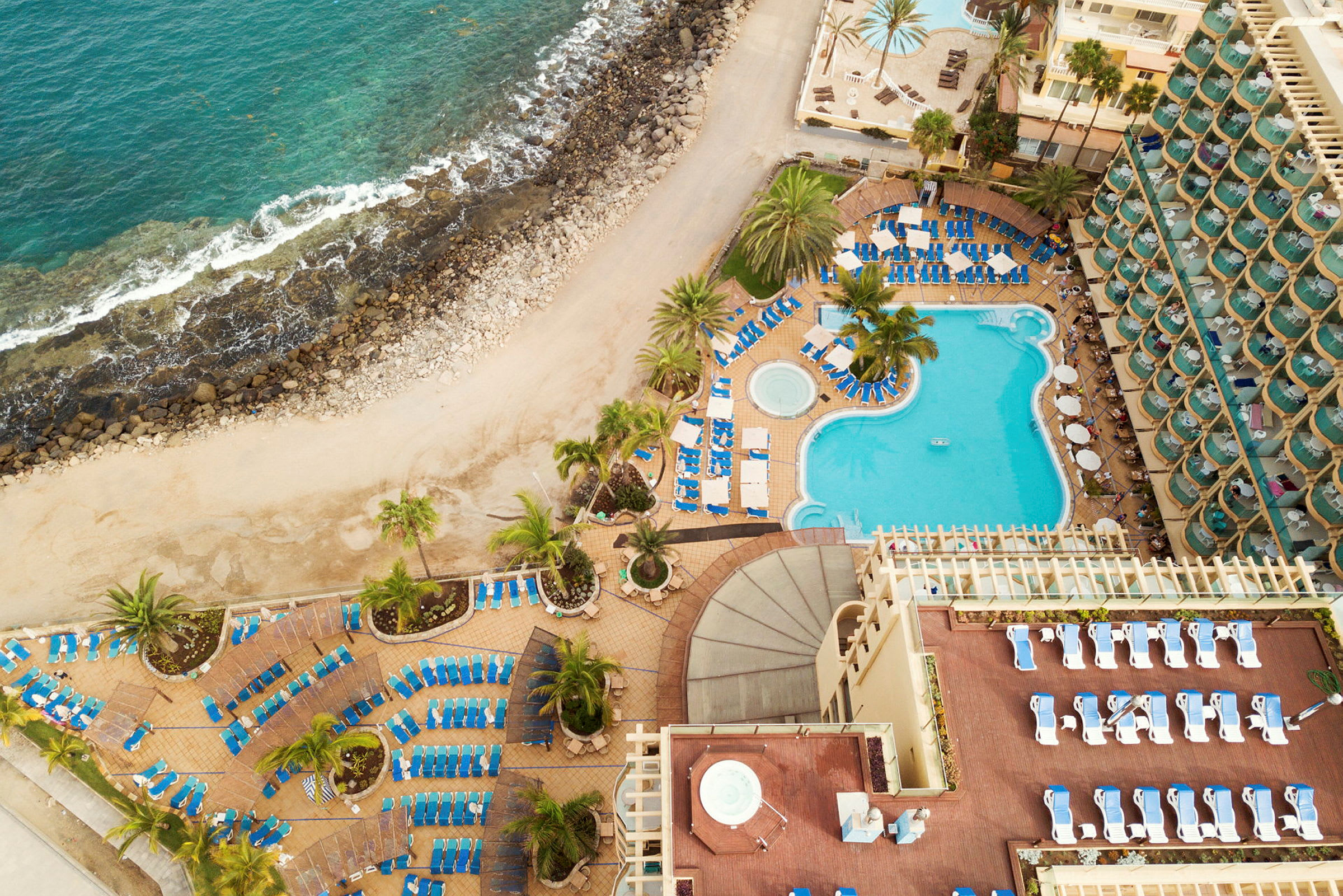 Flygfoto över en hotellpool omgiven av solstolar vid kusten. (AI-genererad text)