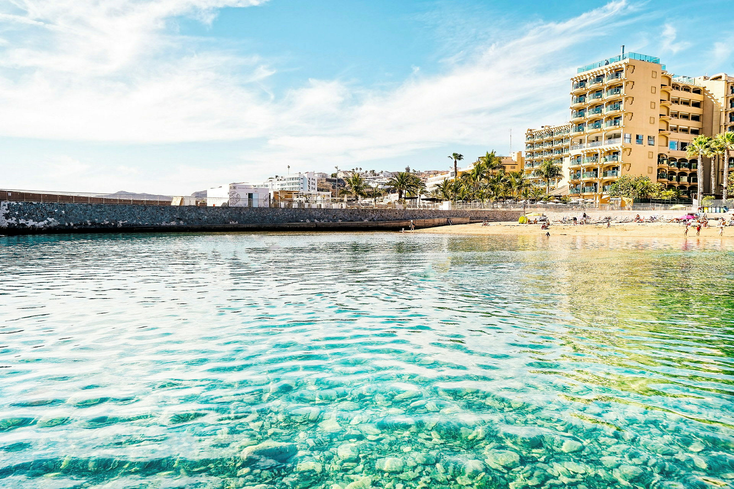 Strand med en klippig havsmur och hotell i bakgrunden. (AI-genererad text)