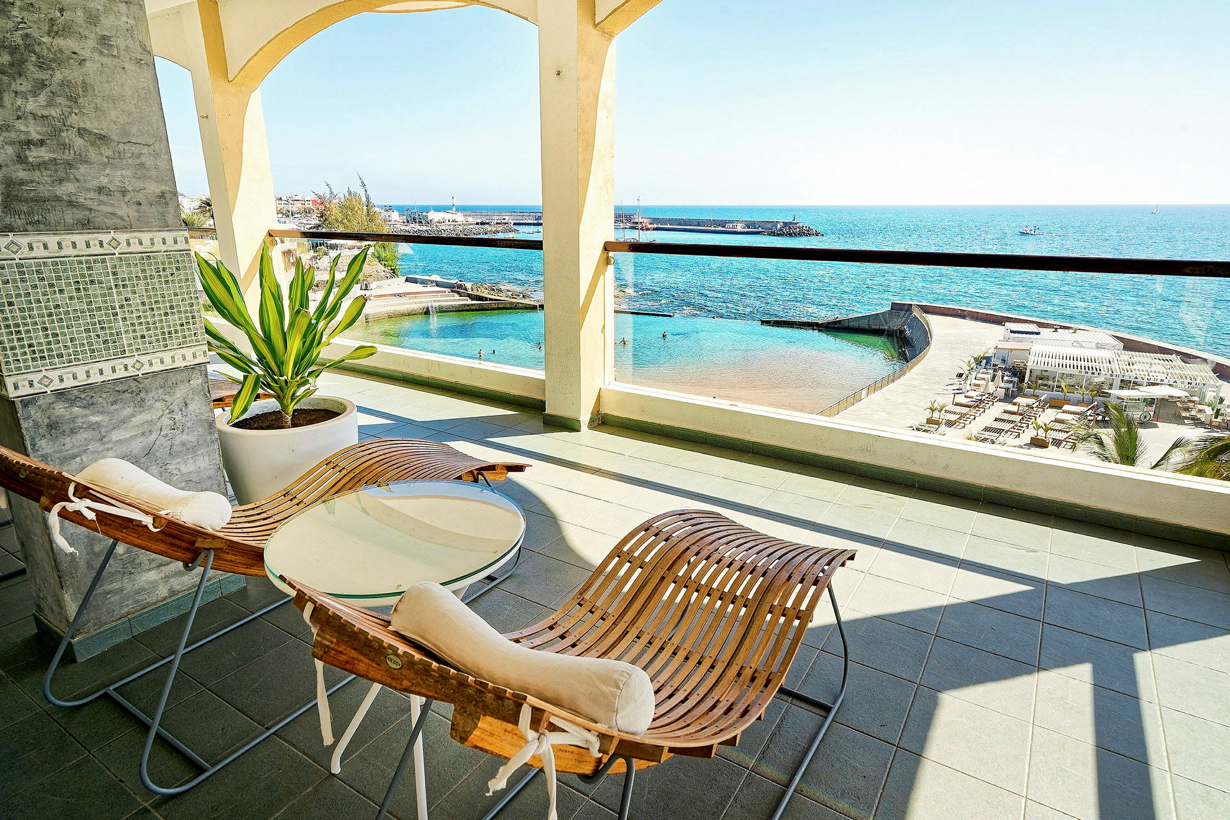 Terrass med två trästolar, ett bord och utsikt över havet och en pool. (AI-genererad text)