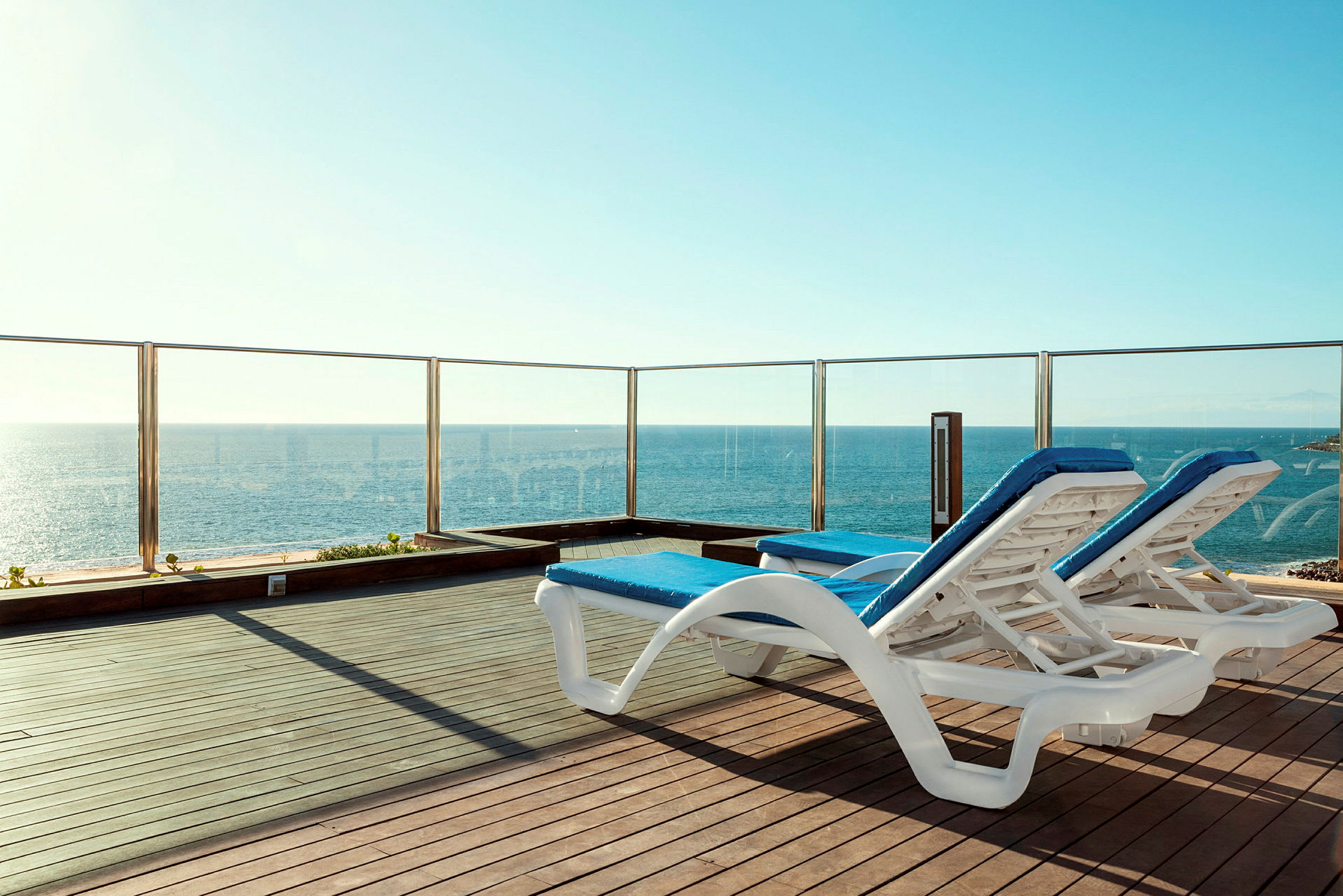 Två solstolar med blå dynor står på en terrass med glasräcke och utsikt över havet. (AI-genererad text)