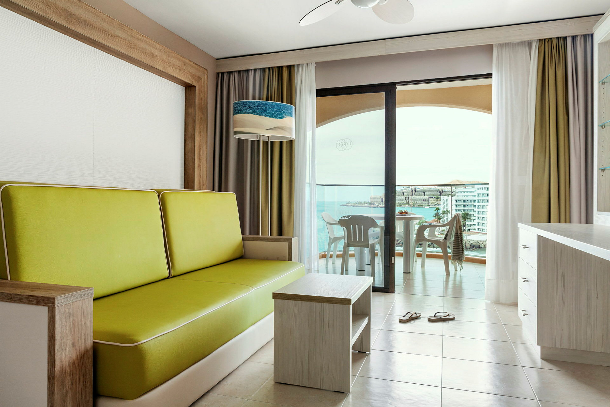 Interiör av ett hotellrum med en grön soffa, ett bord och fönster som vetter mot en balkong med utsikt över havet. (AI-genererad text)