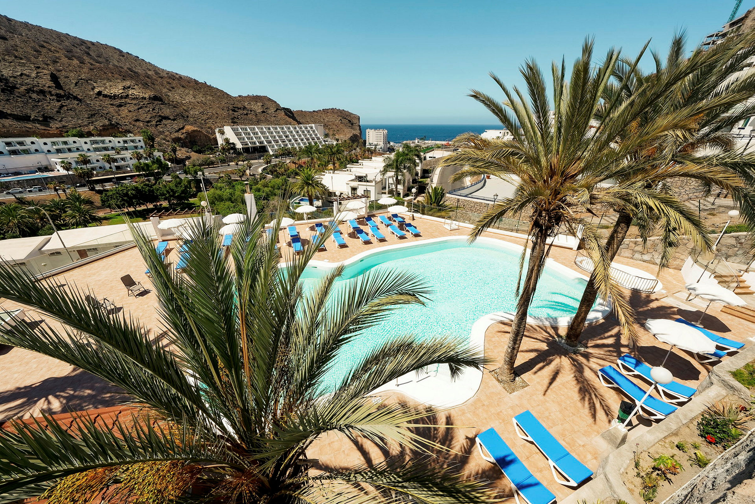 Vy över en hotellpool omgiven av solstolar och palmer med berg och hav i bakgrunden. (AI-genererad text)
