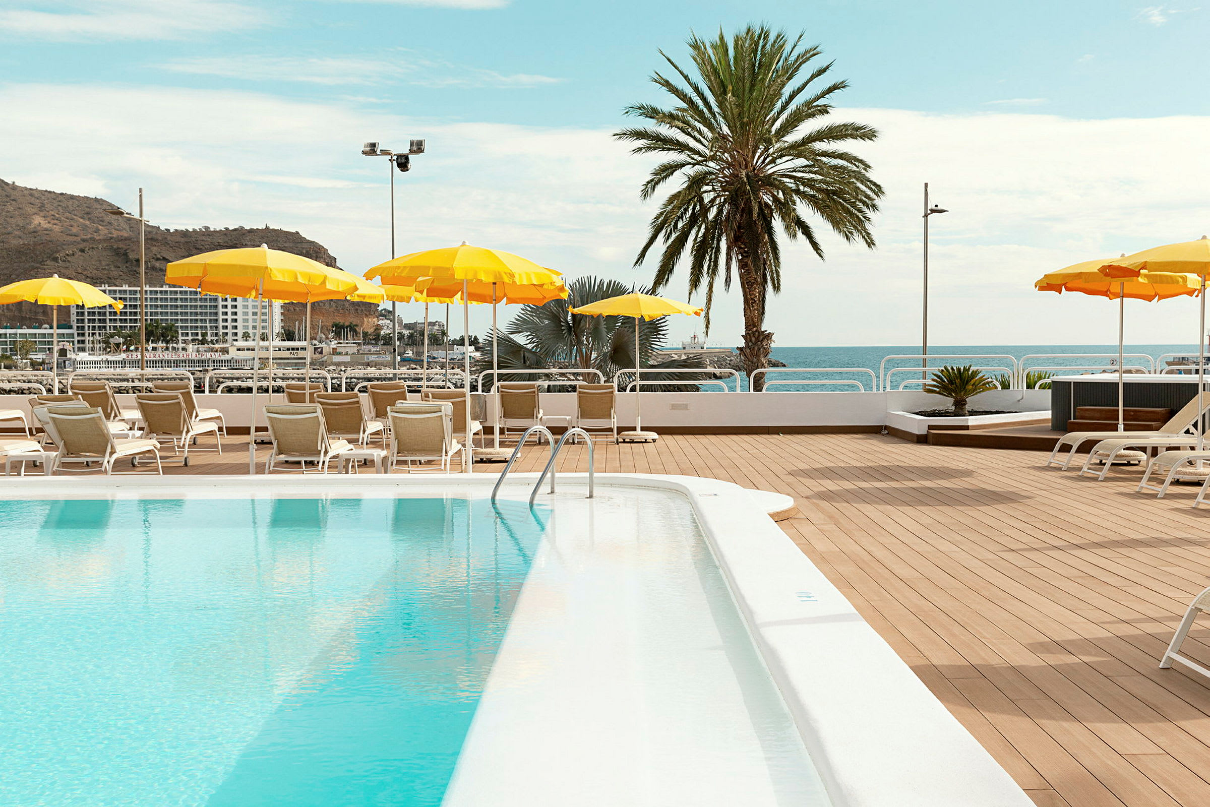En solterrass vid en pool med solstolar och gula parasoller utplacerade. En palm och havet syns i bakgrunden. (AI-genererad text)