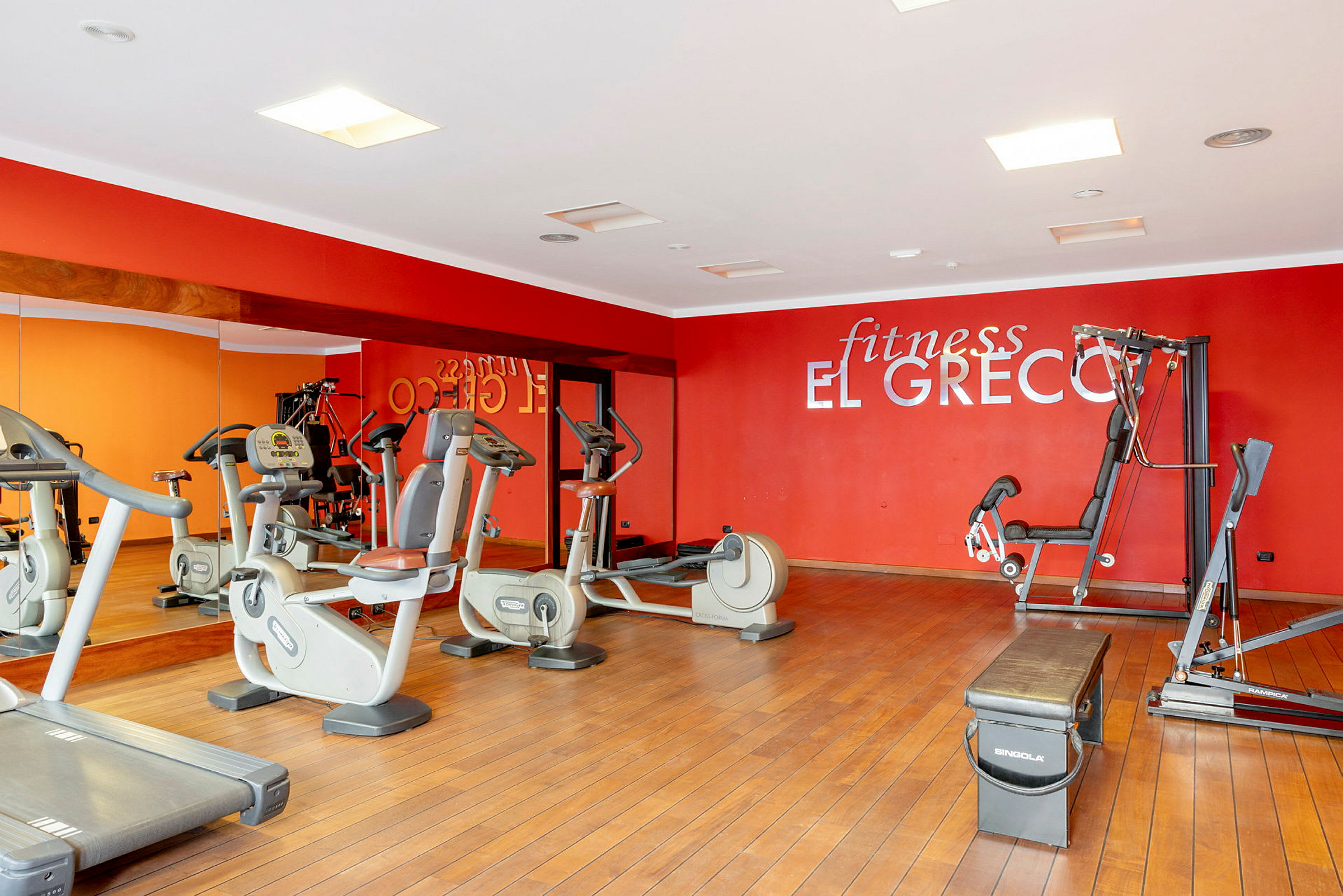 Ett gym med träningsmaskiner, speglar och en röd vägg som har texten 'fitness El Greco'. (AI-genererad text)