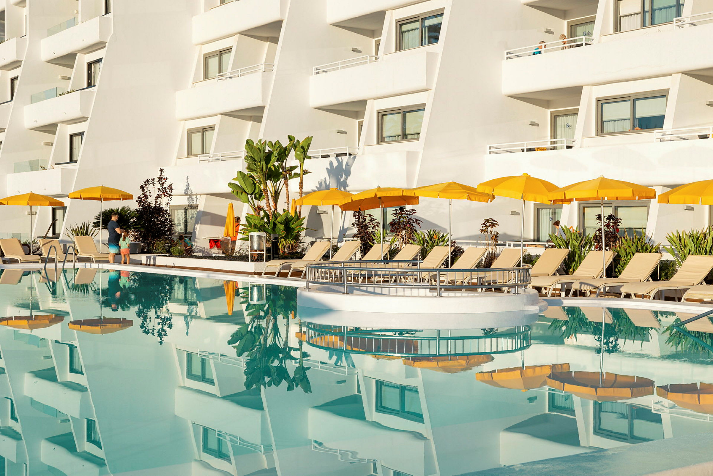 Hotellfasad med balkonger och gulparasol vid en pool. (AI-genererad text)