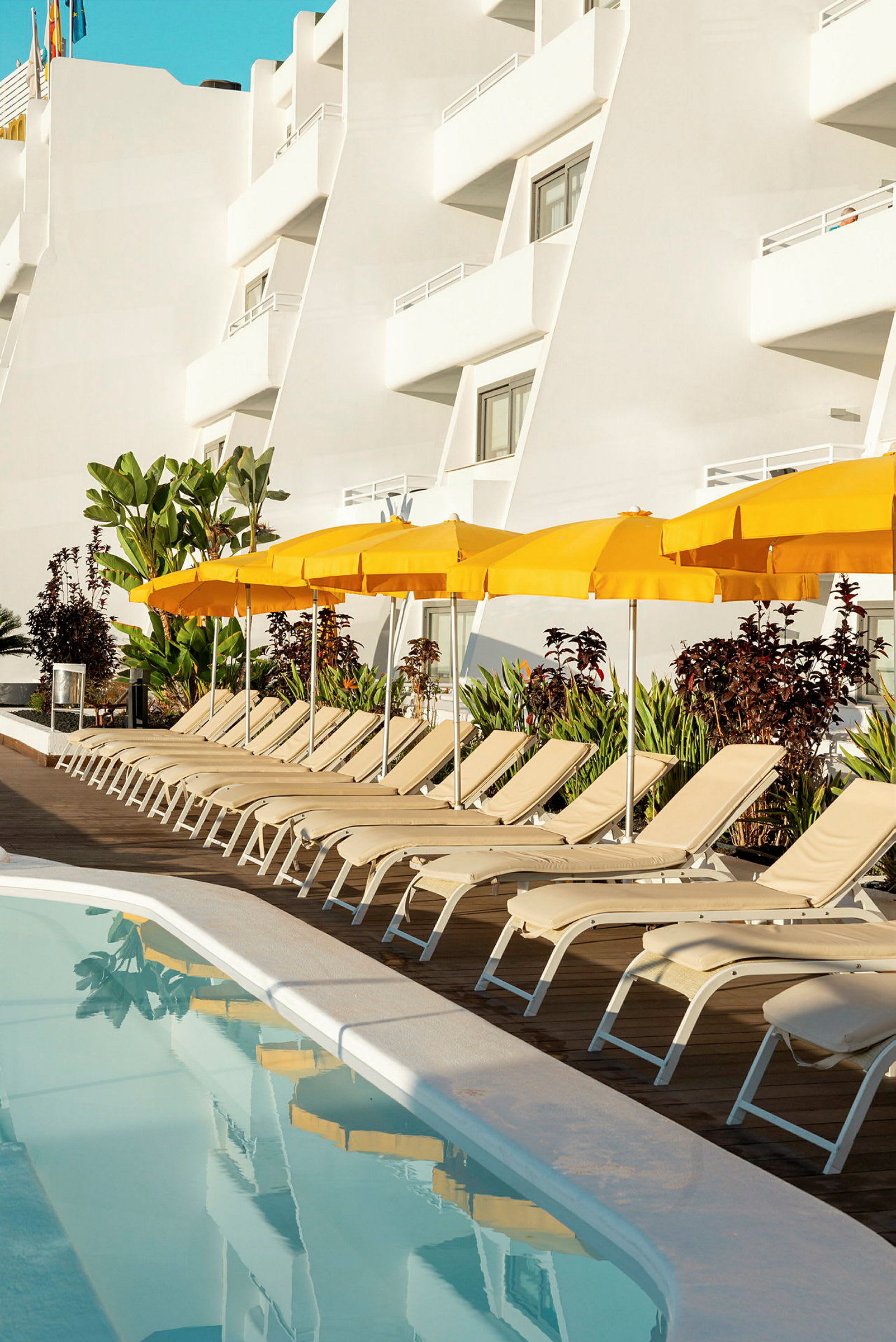 Solstolar bredvid en pool med gula parasoller och en vit byggnad i bakgrunden. (AI-genererad text)
