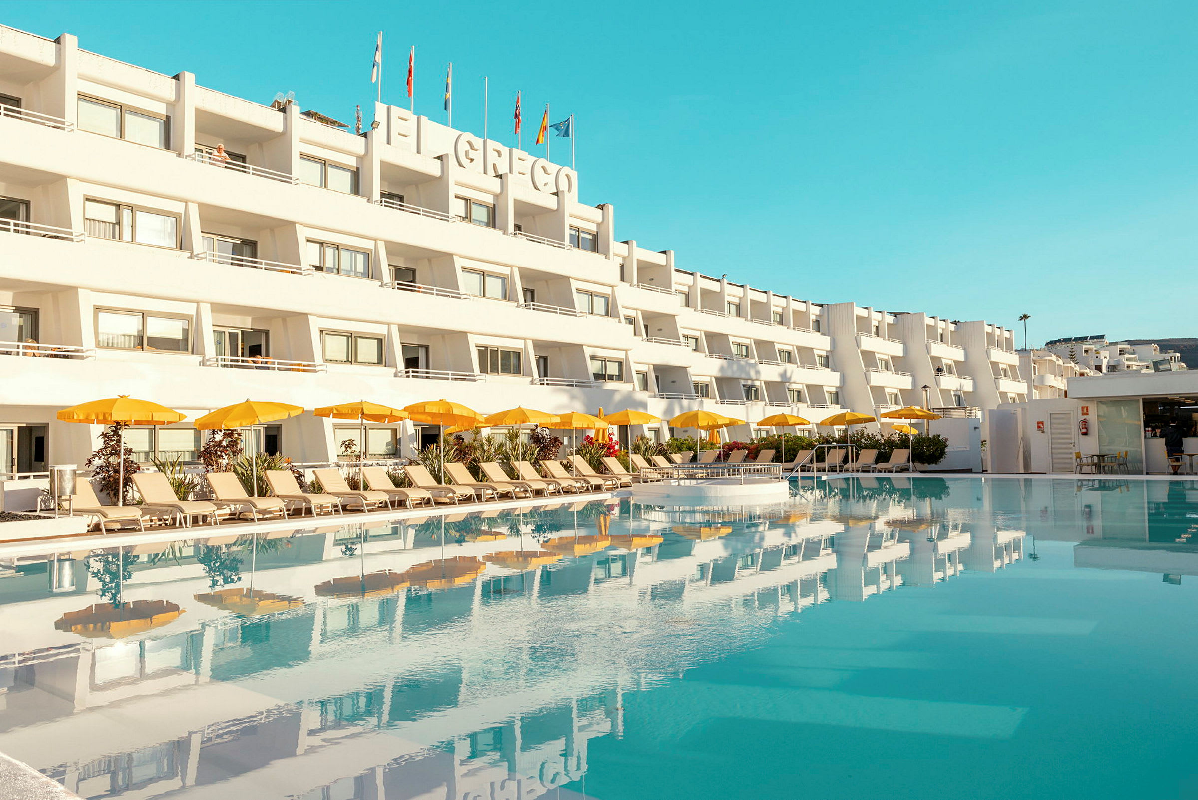 Hotellbyggnad med flera våningar med en pool i förgrunden och solstolar samt gula parasoll. (AI-genererad text)