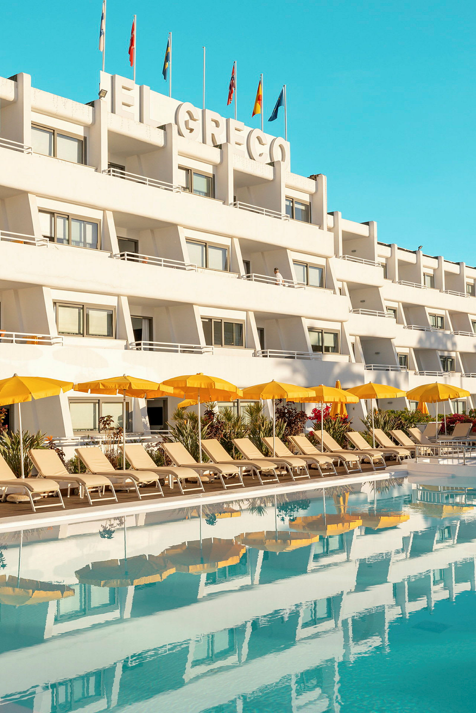 Vit byggnad med balkonger och flaggor på taket, framför en pool med solstolar och gula parasoller. (AI-genererad text)