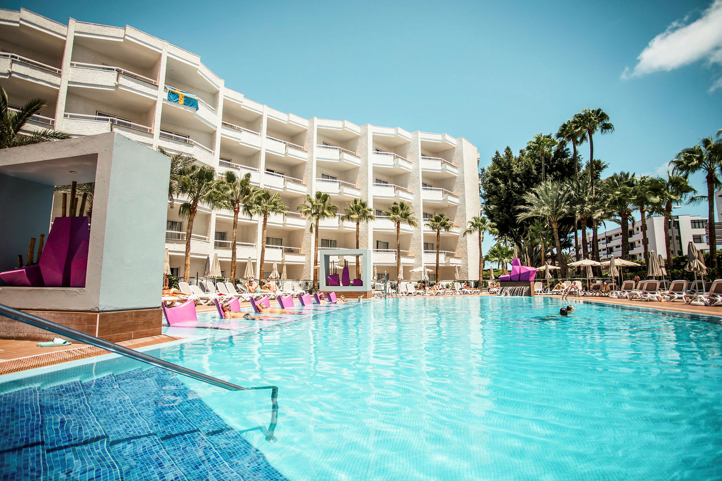 Hotellpool med solstolar och byggnad i bakgrunden. (AI-genererad text)