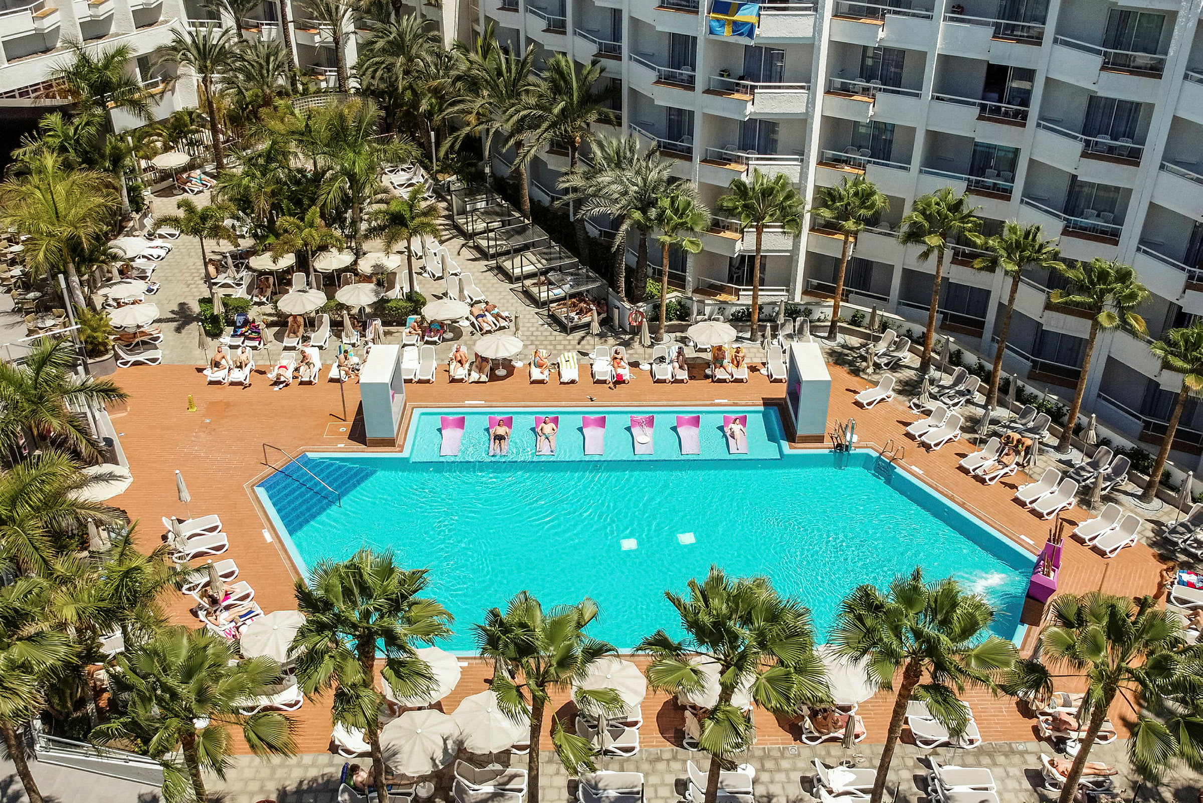 Hotellpool med solstolar och omgivande byggnader. (AI-genererad text)