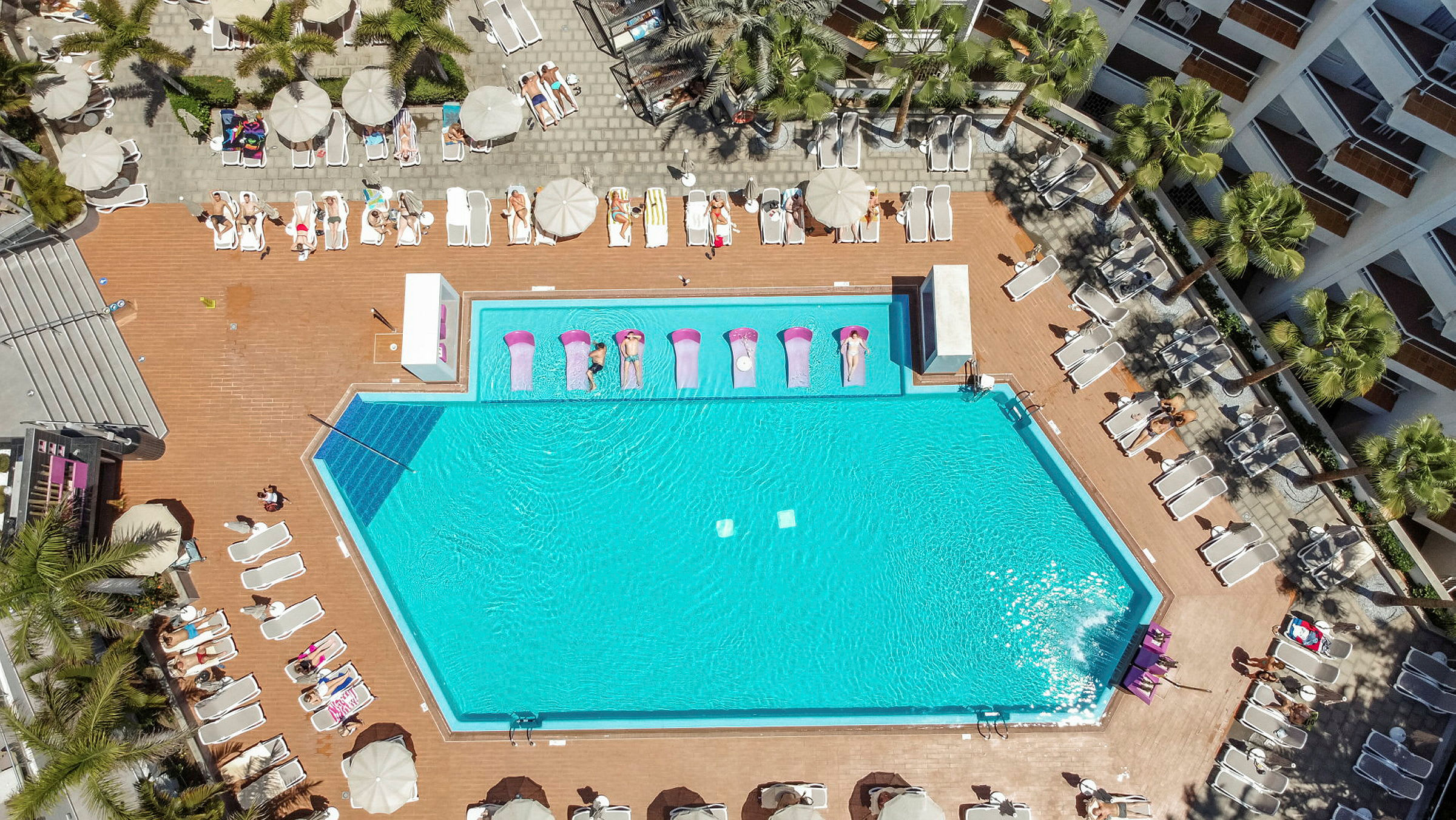 Flygfoto av en swimmingpool omgiven av solstolar och parasoller. (AI-genererad text)