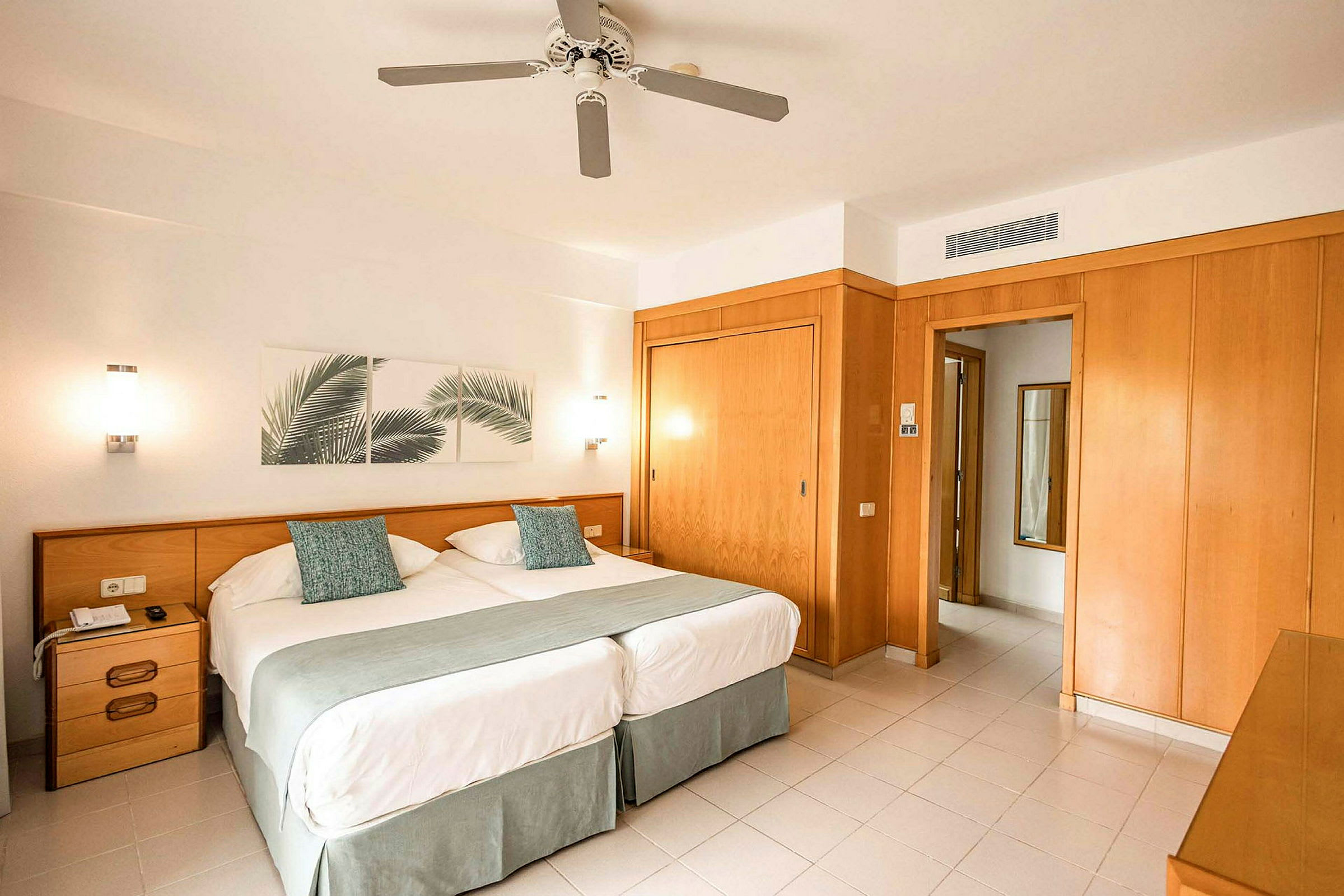 Ett hotellrum med två enkelsängar, väggdekoration med bladmotiv och en takfläkt. (AI-genererad text)