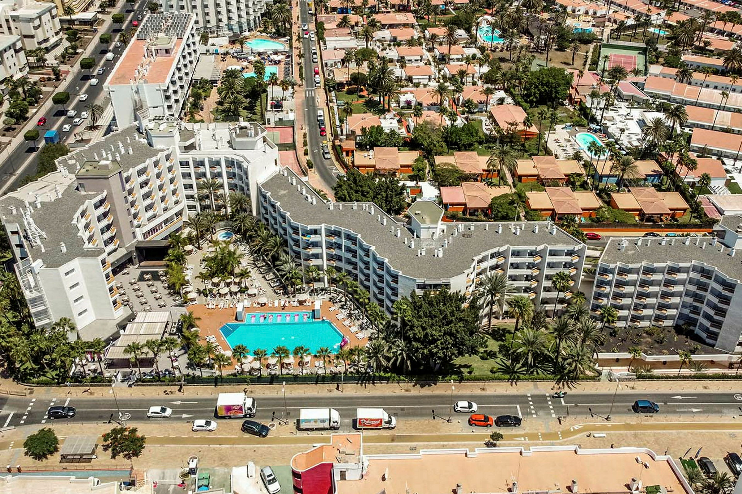 Flygfoto av hotellbyggnader med poolområde, omgivna av träd och mindre hus. (AI-genererad text)