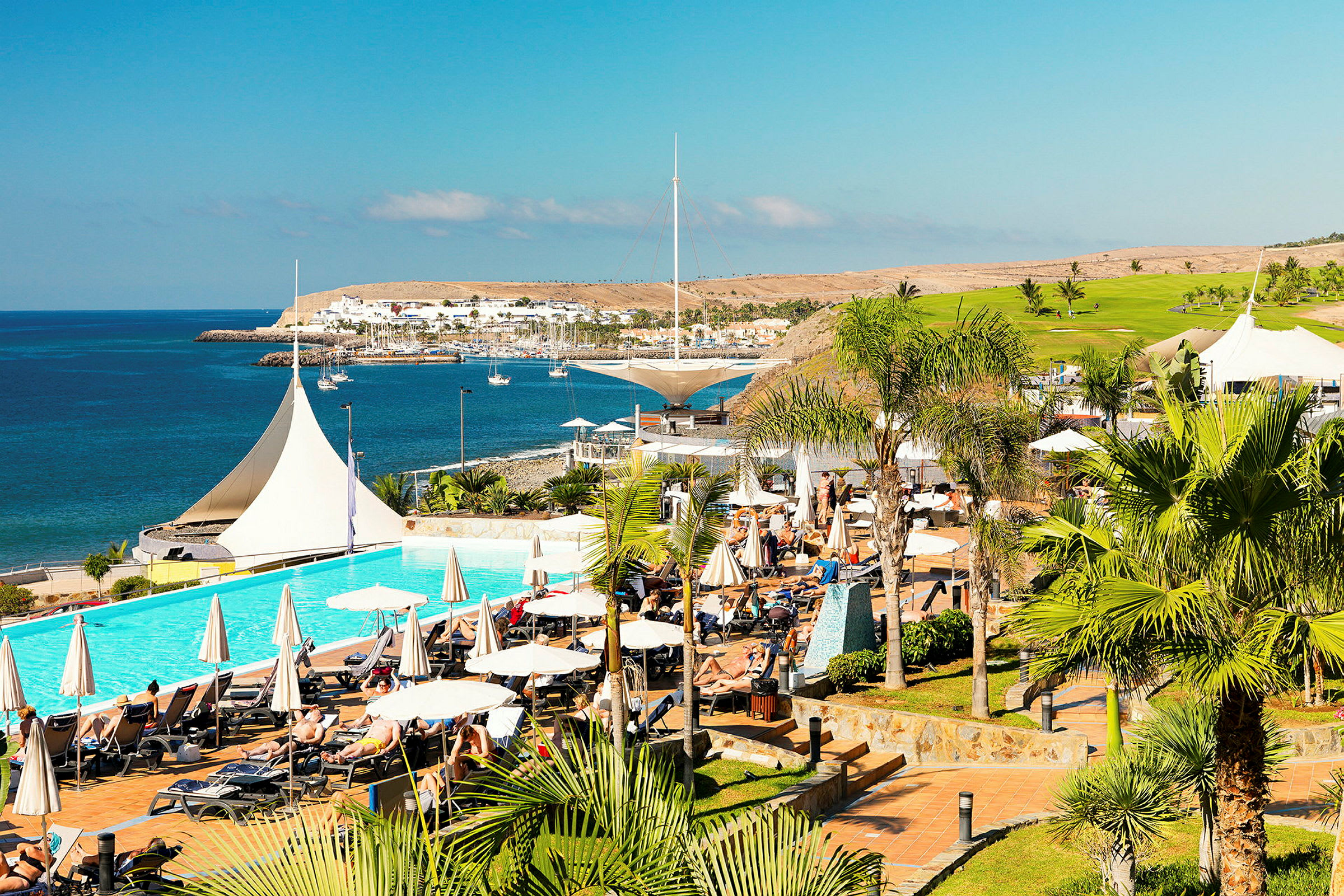 Vy över en resort med pool, solstolar och parasoller, belägen vid kusten med havsutsikt. (AI-genererad text)