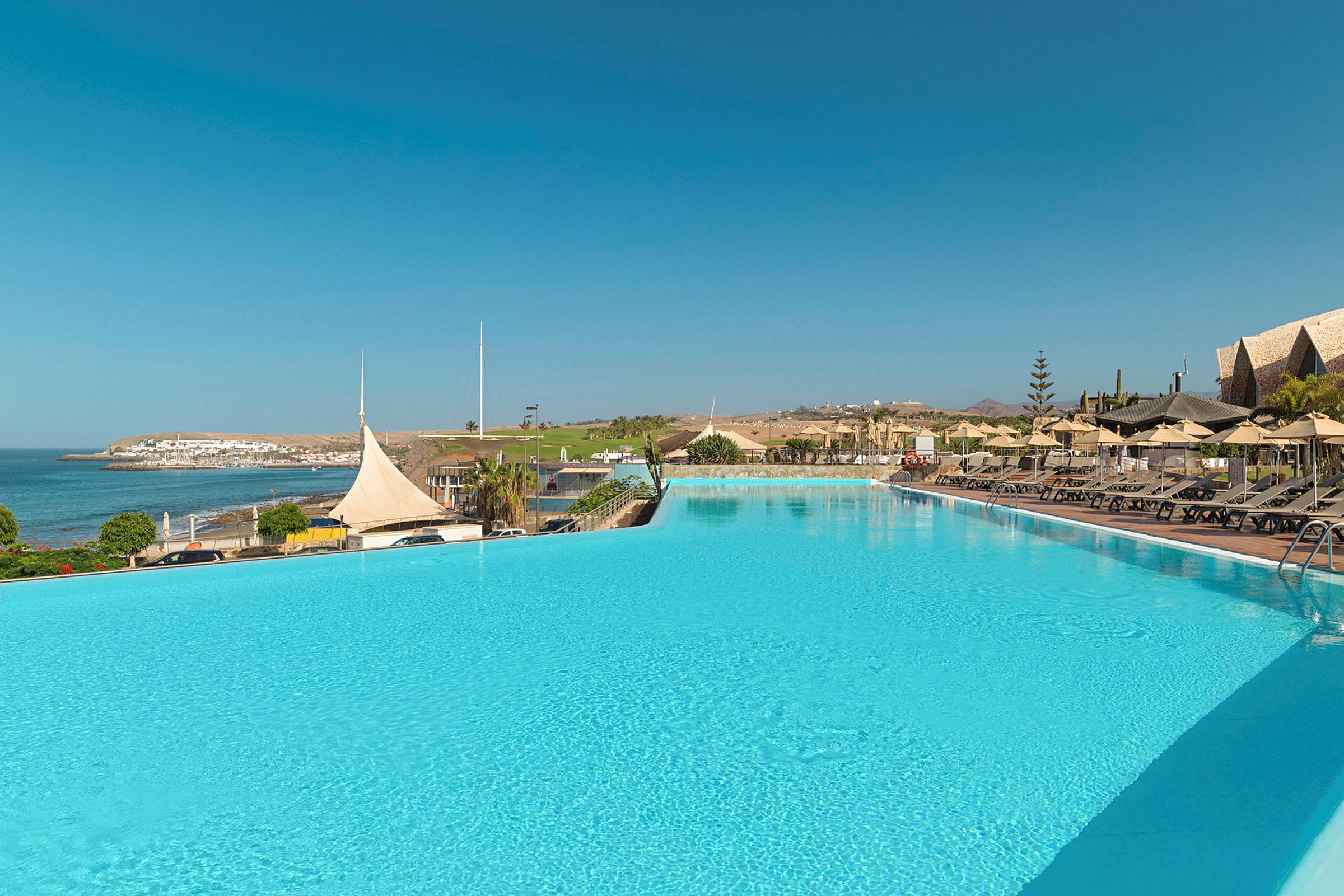En stor infinitypool med utsikt över havet och en kustlinje med vita byggnader. Solstolar och parasoller är placerade längs poolkanten. (AI-genererad text)