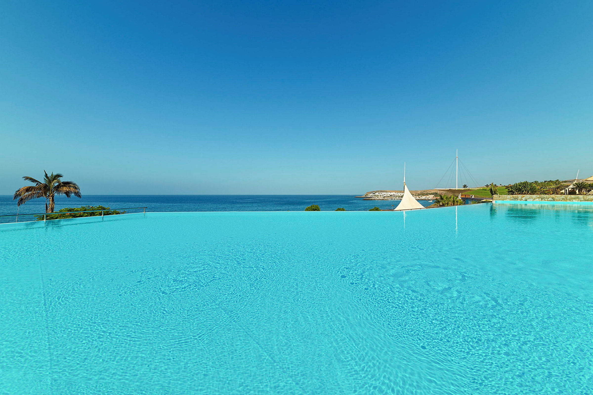 En vid pool med klart vatten och utsikt över havet, med palmer och byggnader i bakgrunden. (AI-genererad text)