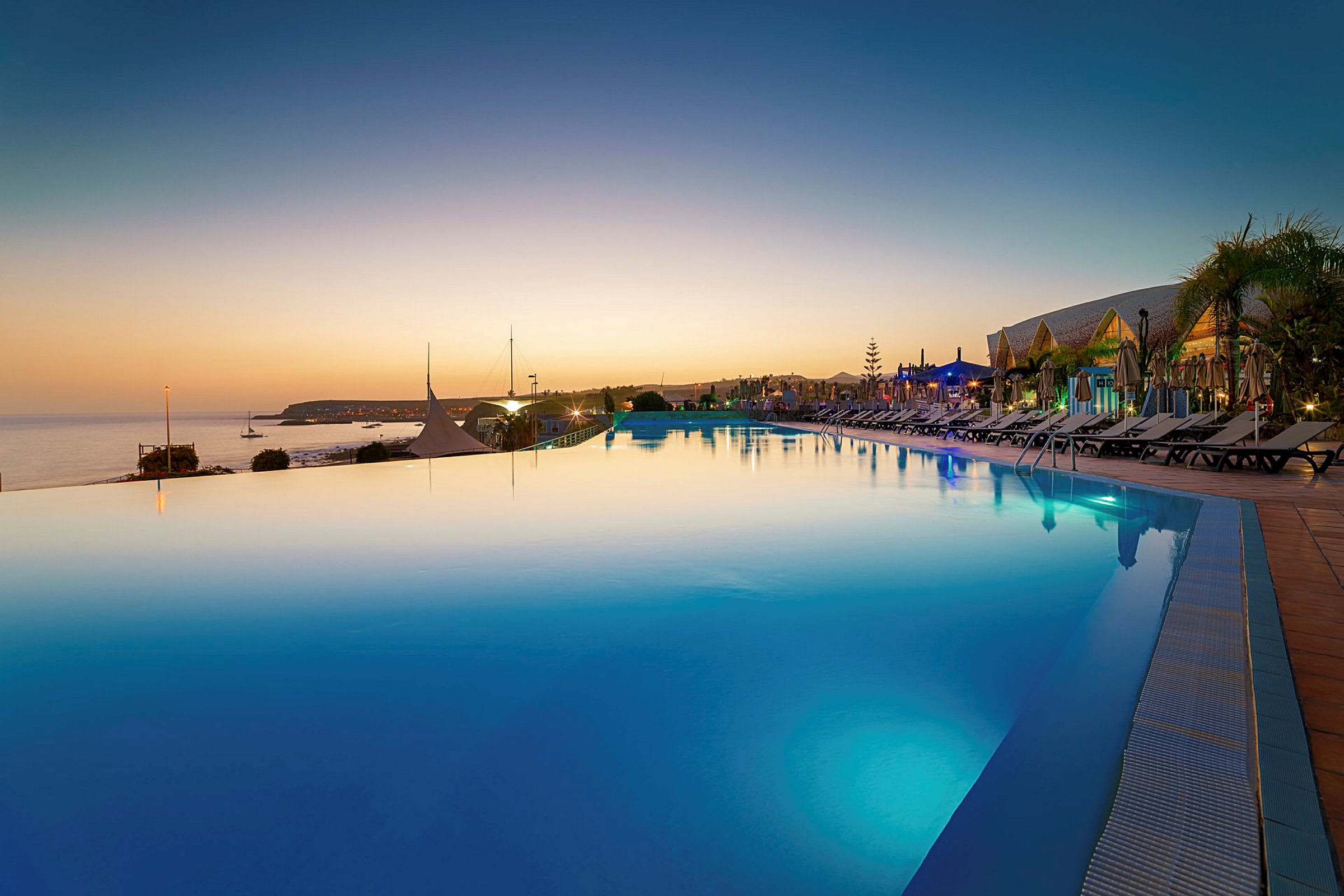 En infinitypool med utsikt över havet vid solnedgången. Solstolar står längs poolkanten. (AI-genererad text)