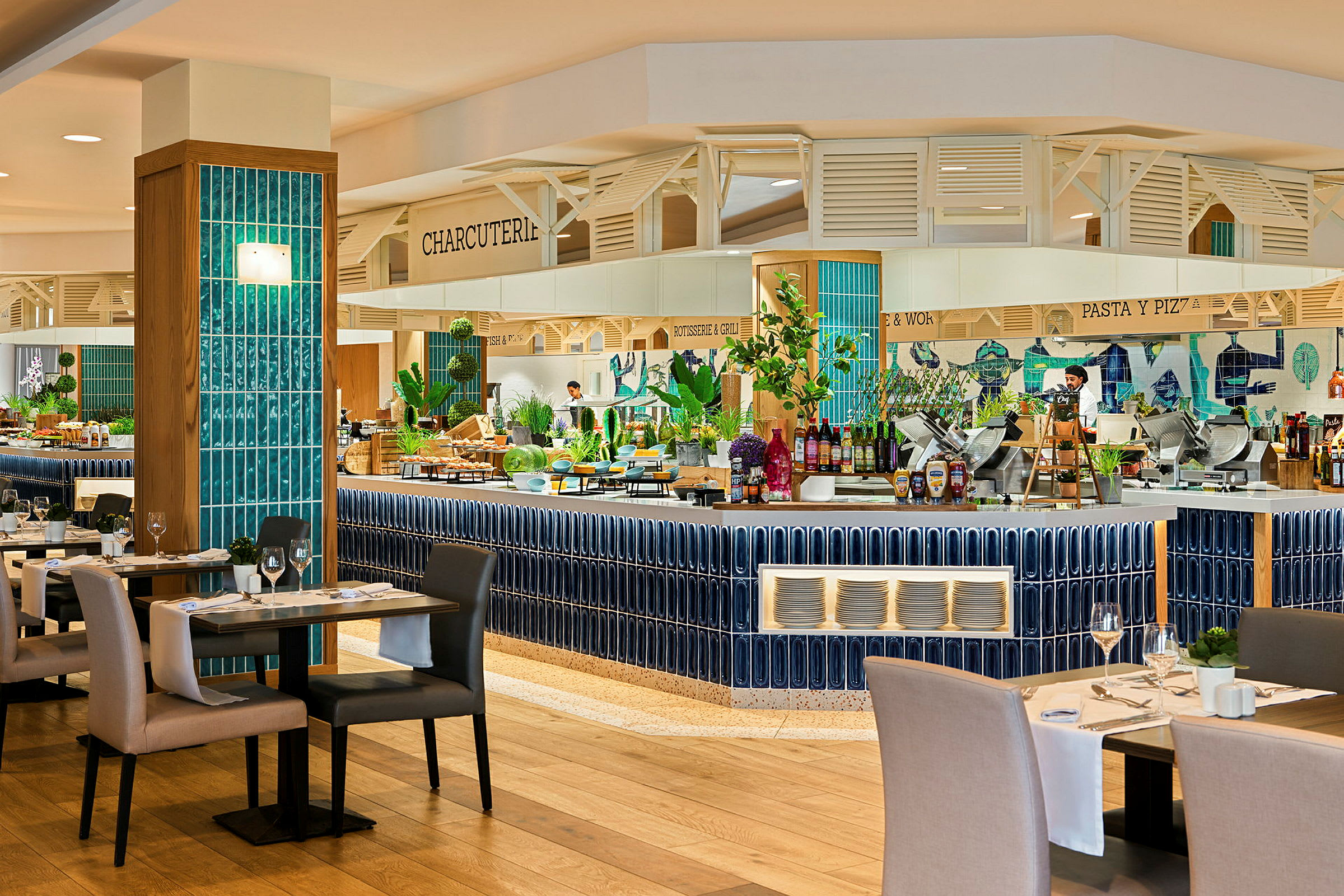 En restaurang med uppdukade bord och ett öppet köksområde. Ytorna är dekorerade med blå kakel och färgglada detaljer. (AI-genererad text)