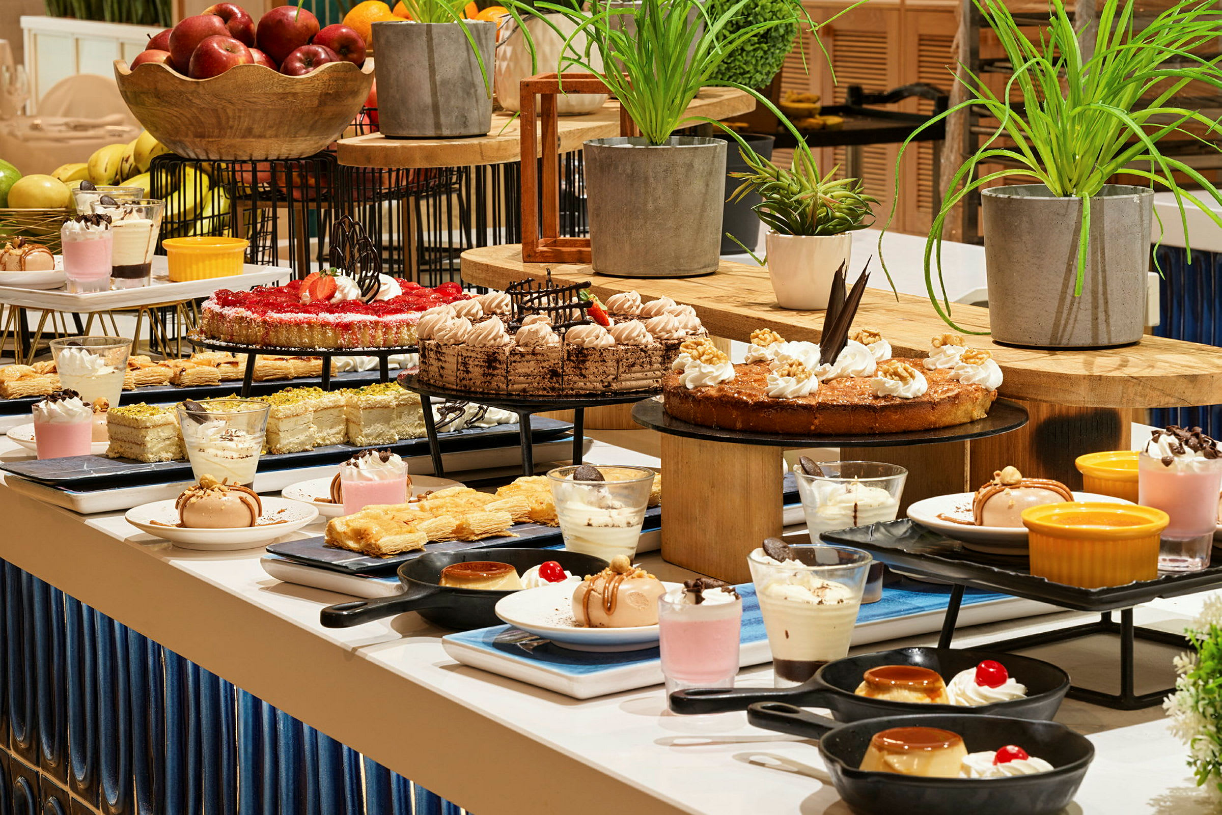 En buffé med olika desserter, inklusive kakor, bakverk, mousse och frukter. Växter är placerade som dekoration. (AI-genererad text)