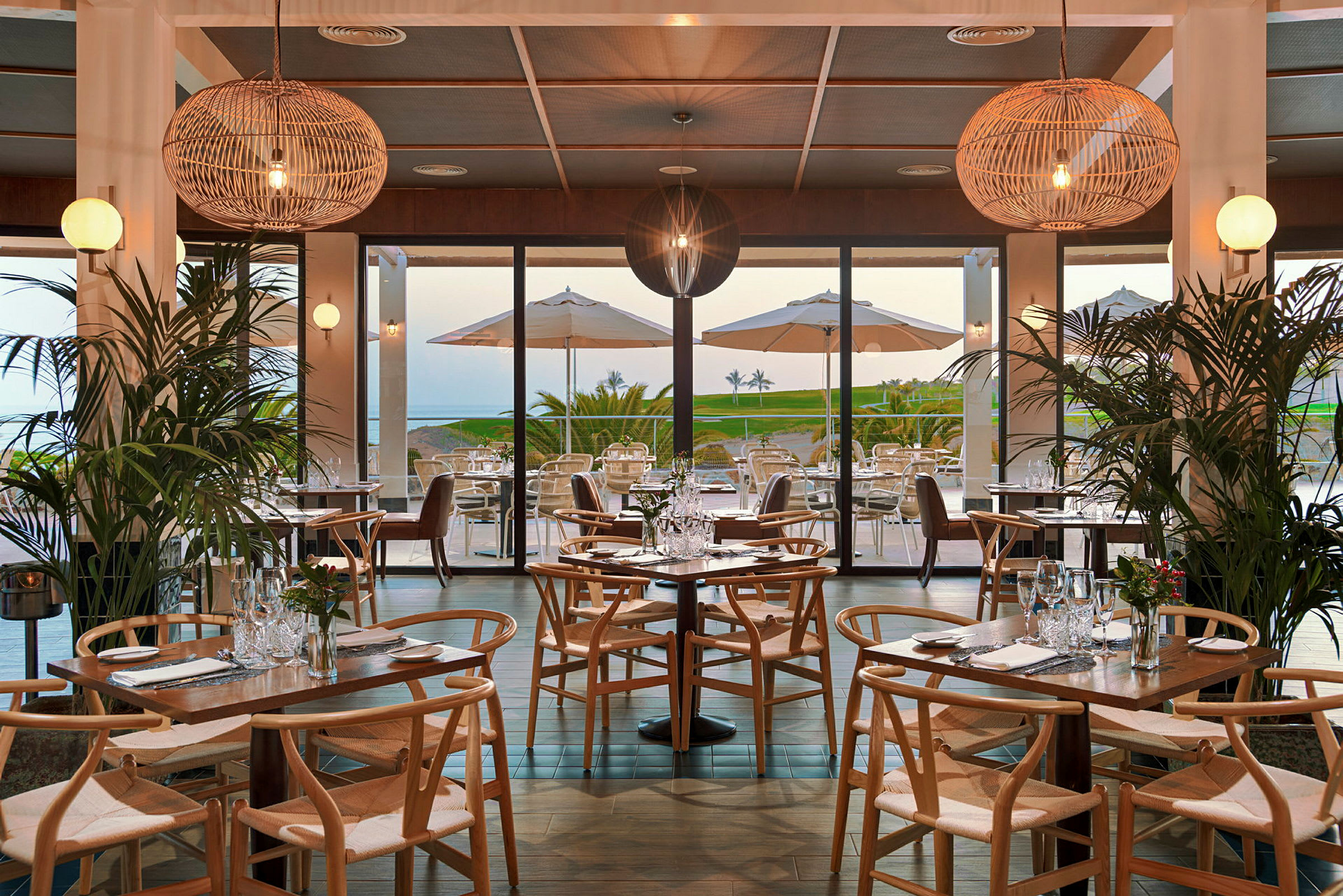 En restaurang med träbord och stolar, dekorerad med växter och stora hängande lampor. I bakgrunden syns en terrass med parasoller och en utsikt mot ett grönområde och havet. (AI-genererad text)