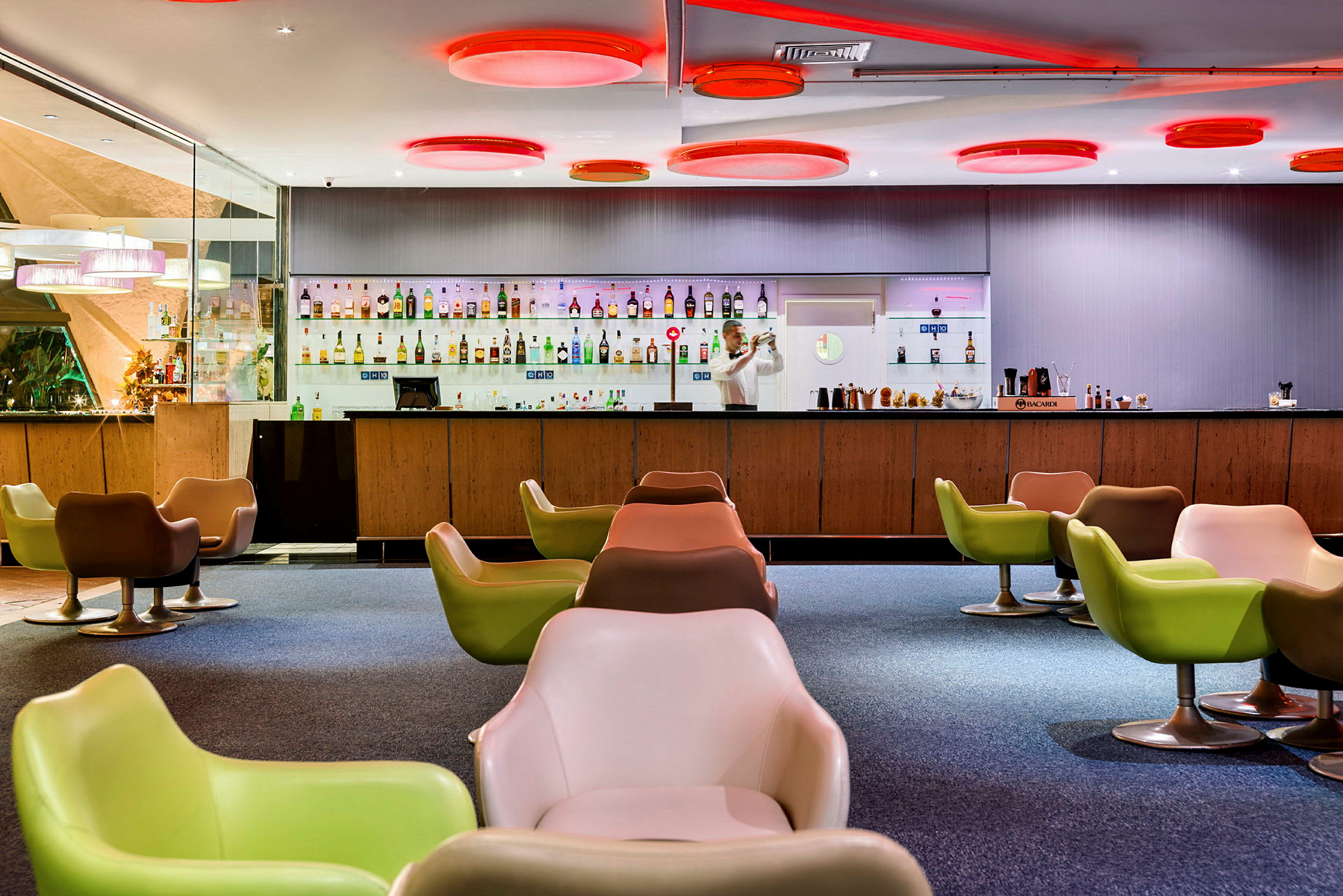 En modern bar med färgglada stolar, en bartender som förbereder drinkar bakom disken och flaskor uppställda prydligt på hyllor. (AI-genererad text)