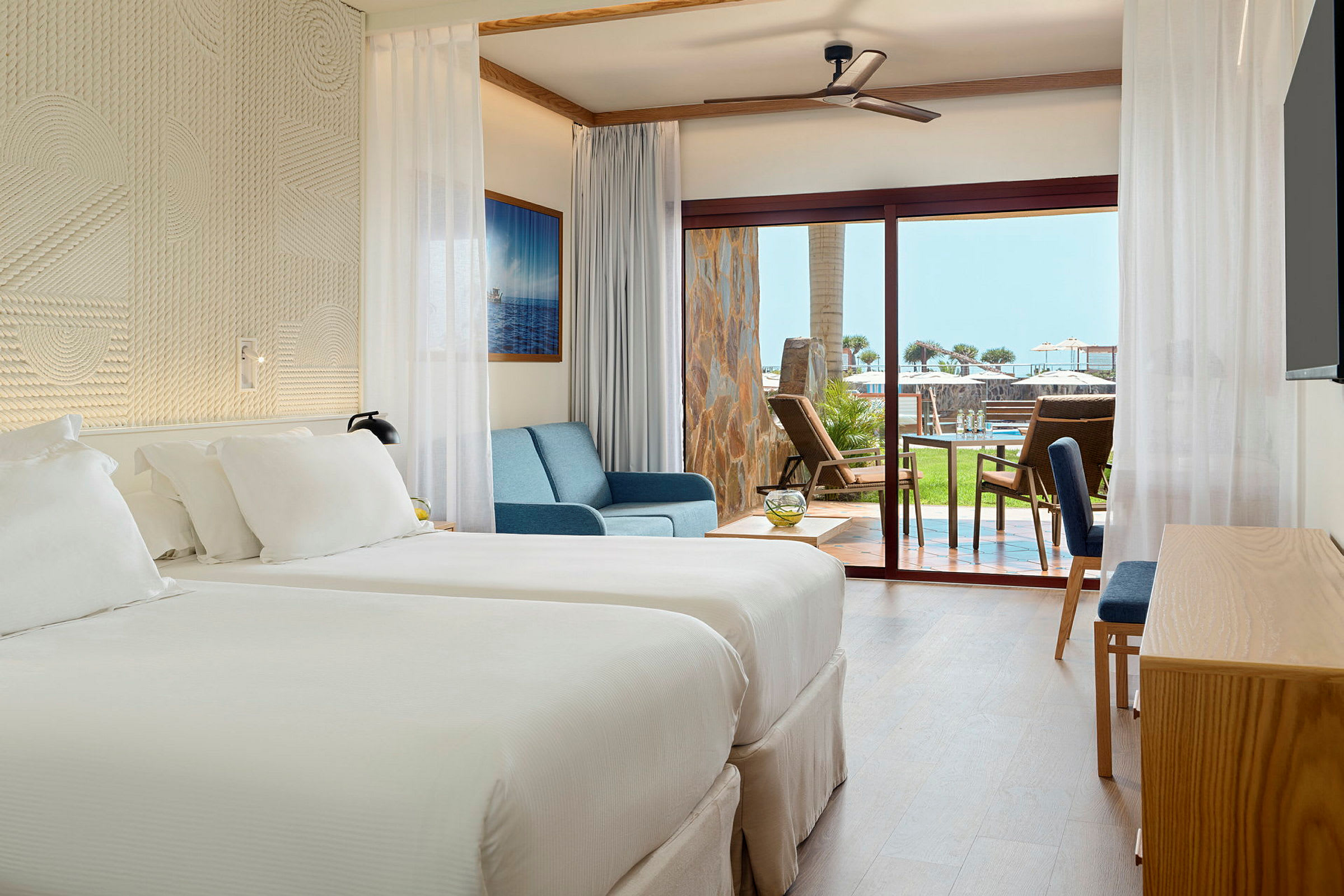 Ett hotellrum med två sängar, blå soffa och stora glasdörrar som leder till en terrass med utsikt över gräsmatta och poolområde. (AI-genererad text)