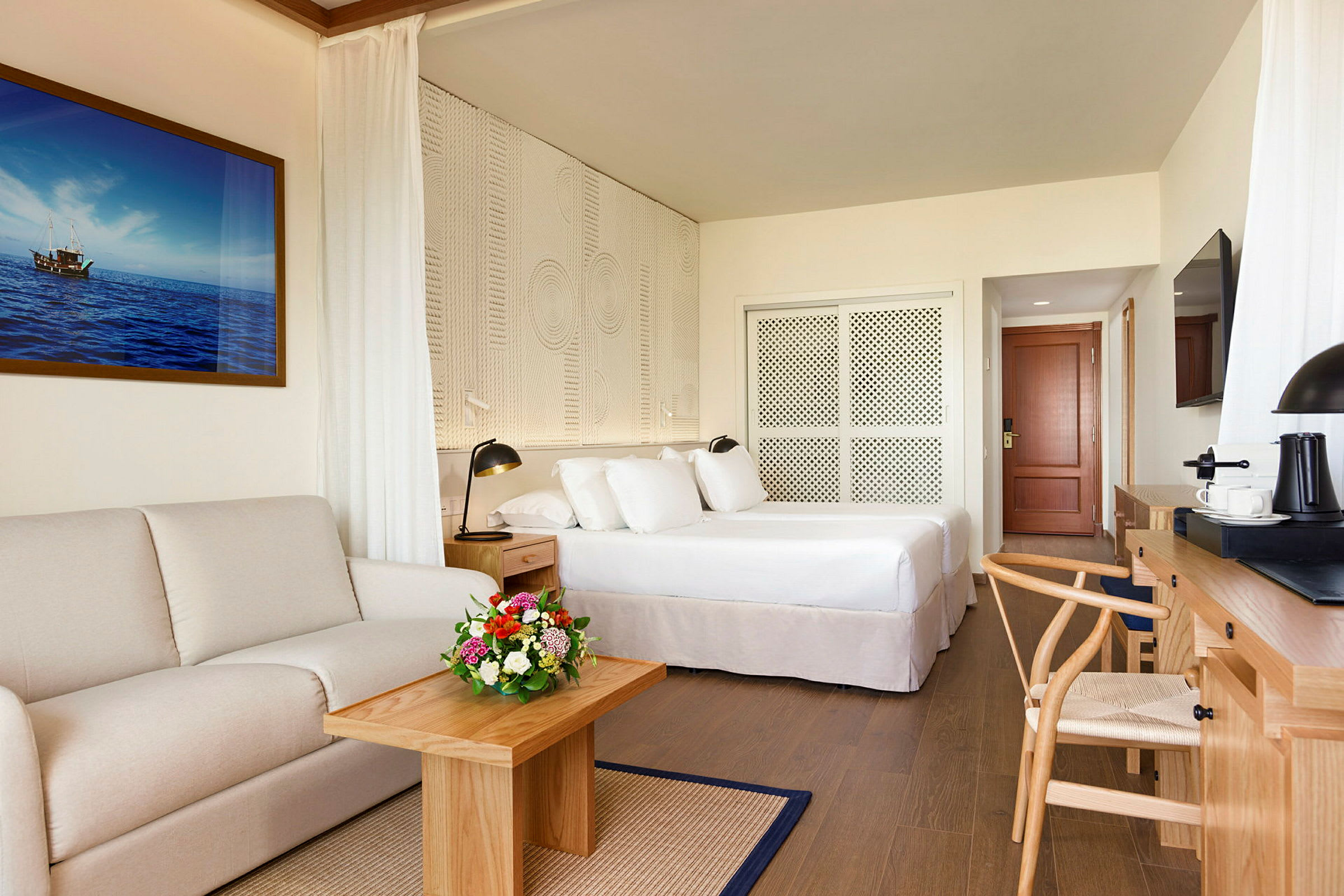 Ett hotellrum med en mjuk soffa, ett bord med en blomsterbukett, en säng med vita sängkläder, en skrivbordshörna och en väggmålning av havet. (AI-genererad text)