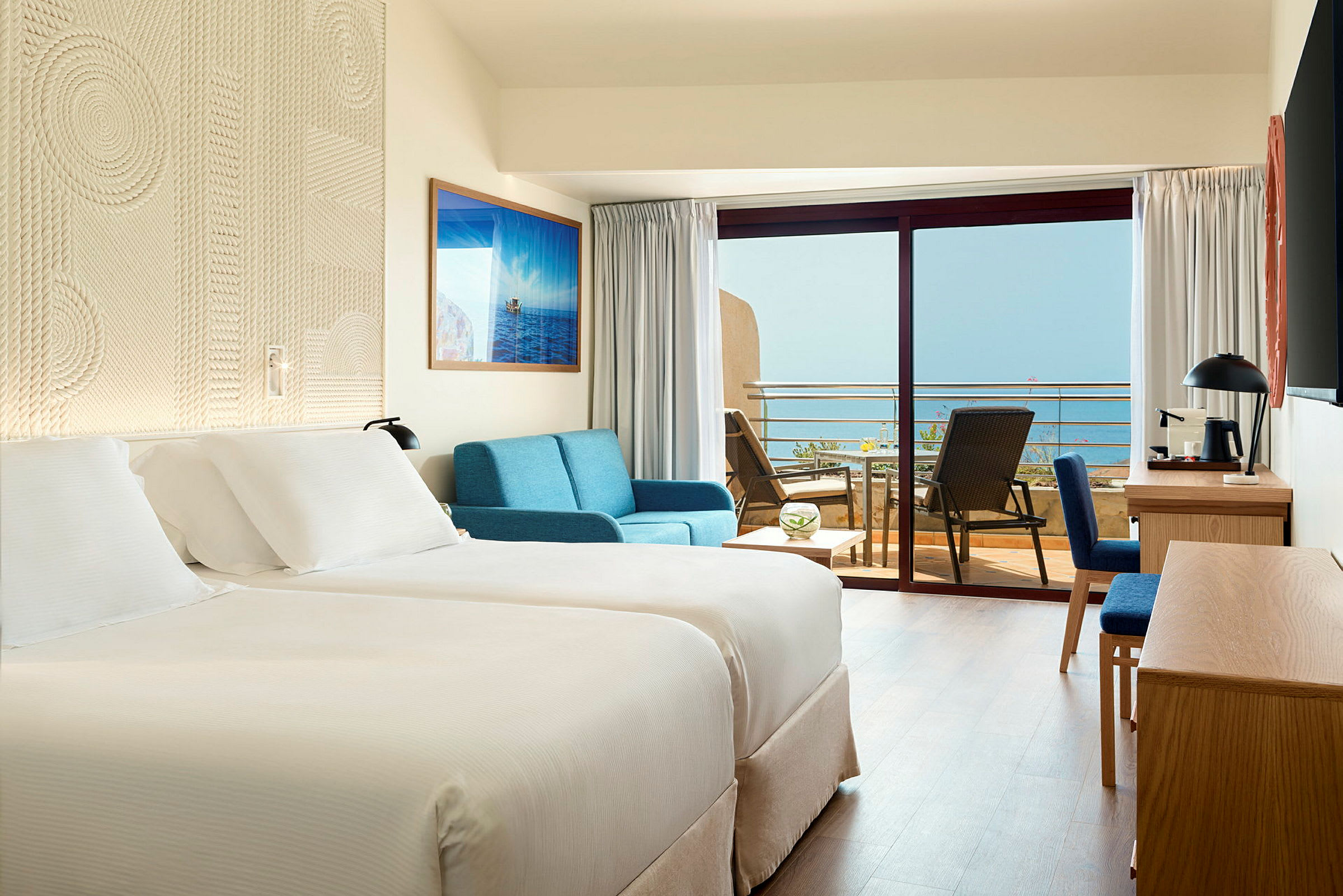 Ett hotellrum med två enkelsängar, en soffa, ett skrivbord och en balkong med utsikt över havet. (AI-genererad text)