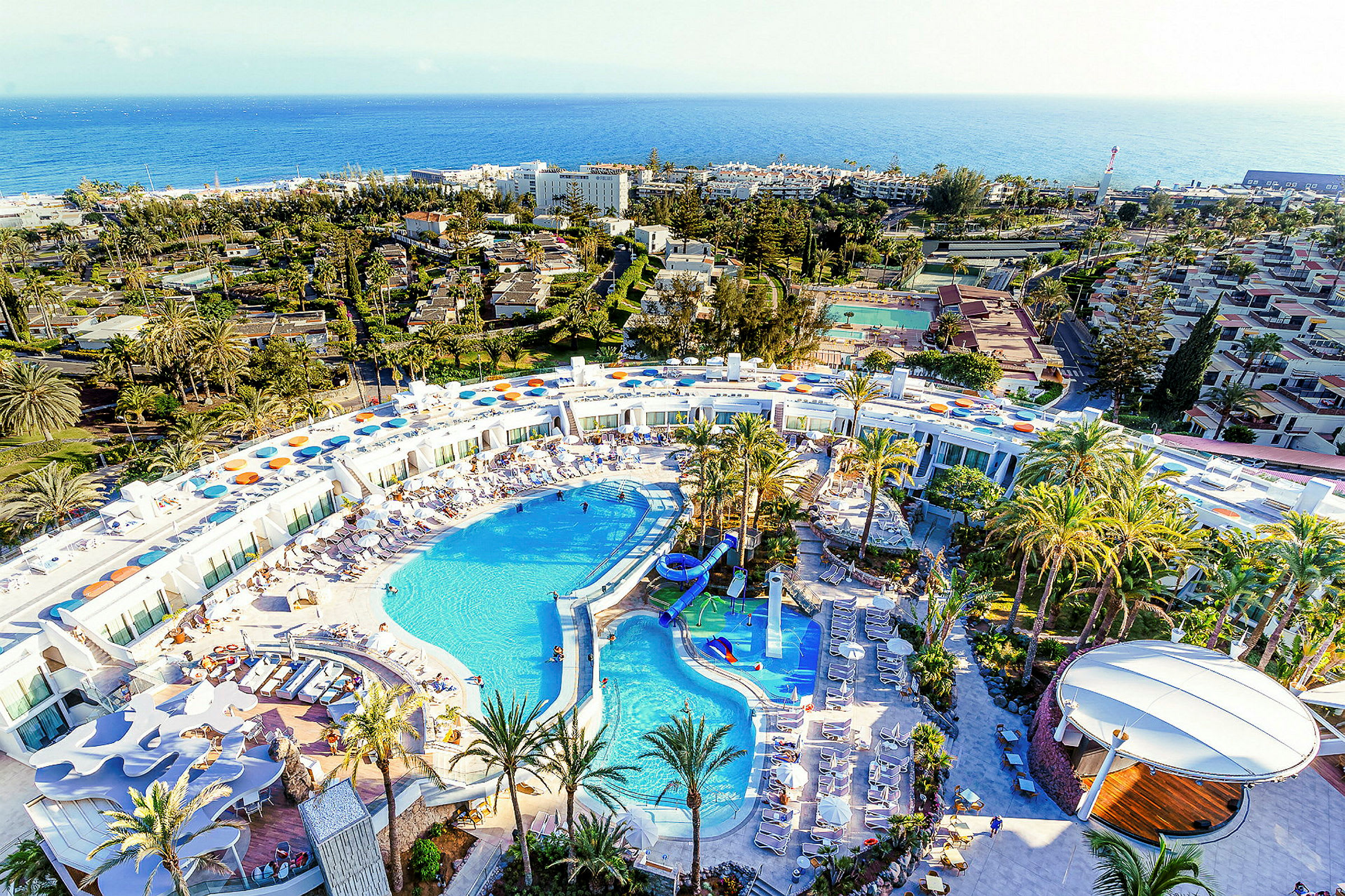 Flygbild över en stor resort med poolområde och omgivet av palmer. (AI-genererad text)