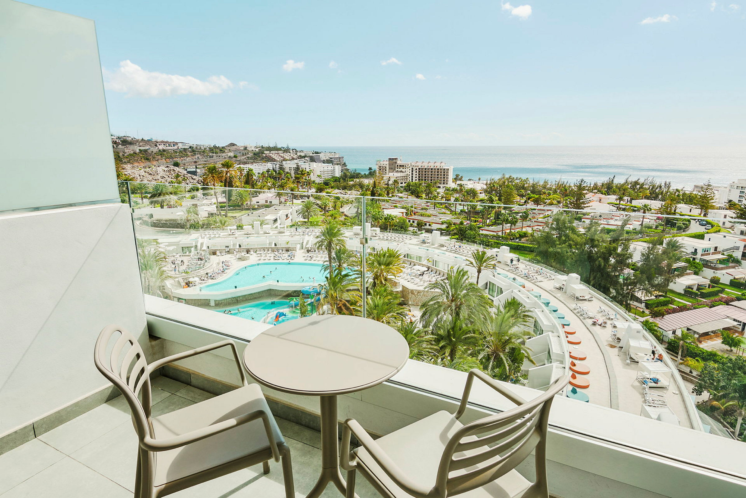 Balkong med två stolar och ett runt bord samt utsikt över hotellområde med pool och hav. (AI-genererad text)