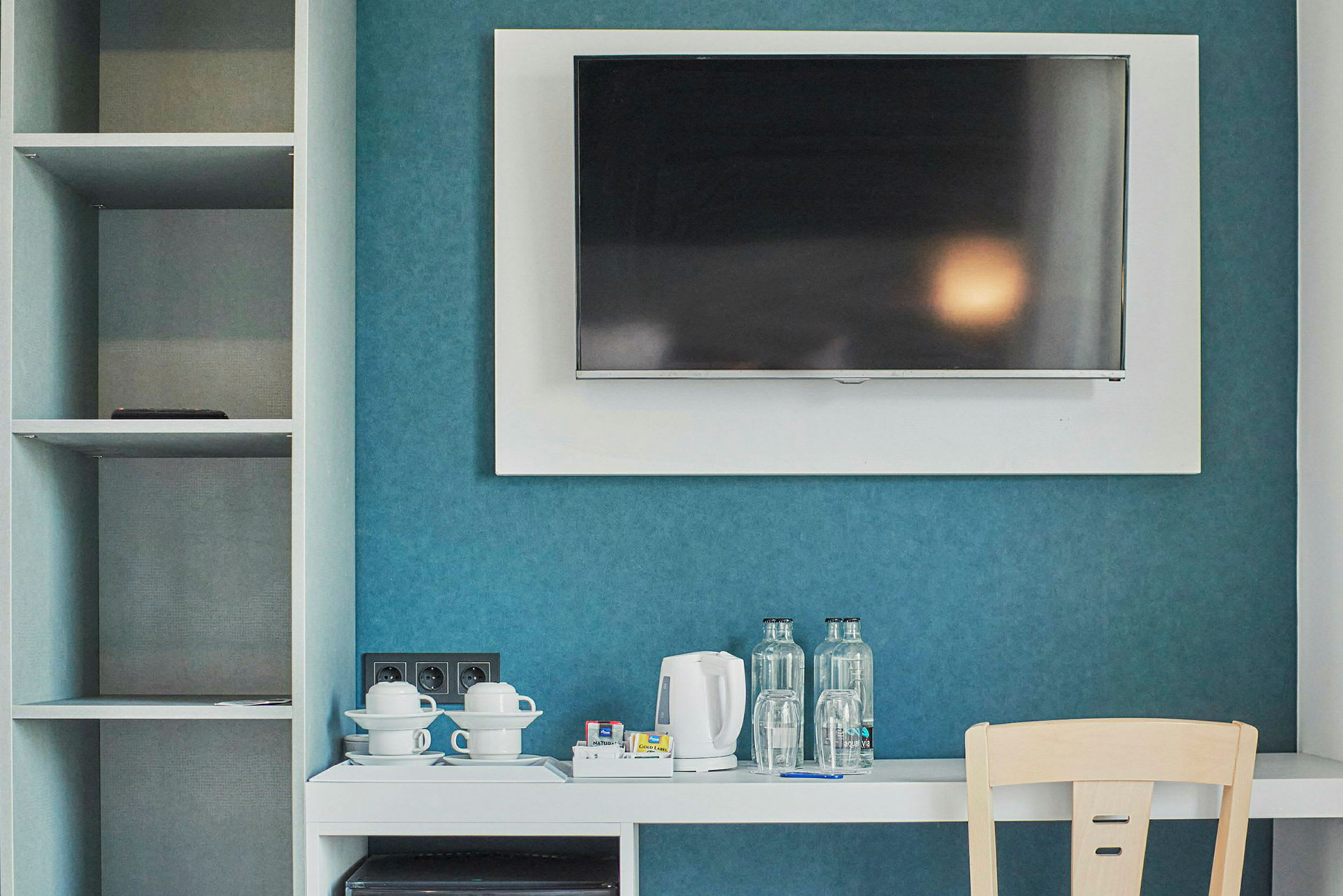 Ett hotellrum med en väggmonterad TV, en vit möbel med vattenkokare, koppar och flaskor, samt ett öppet skåp på sidan. (AI-genererad text)