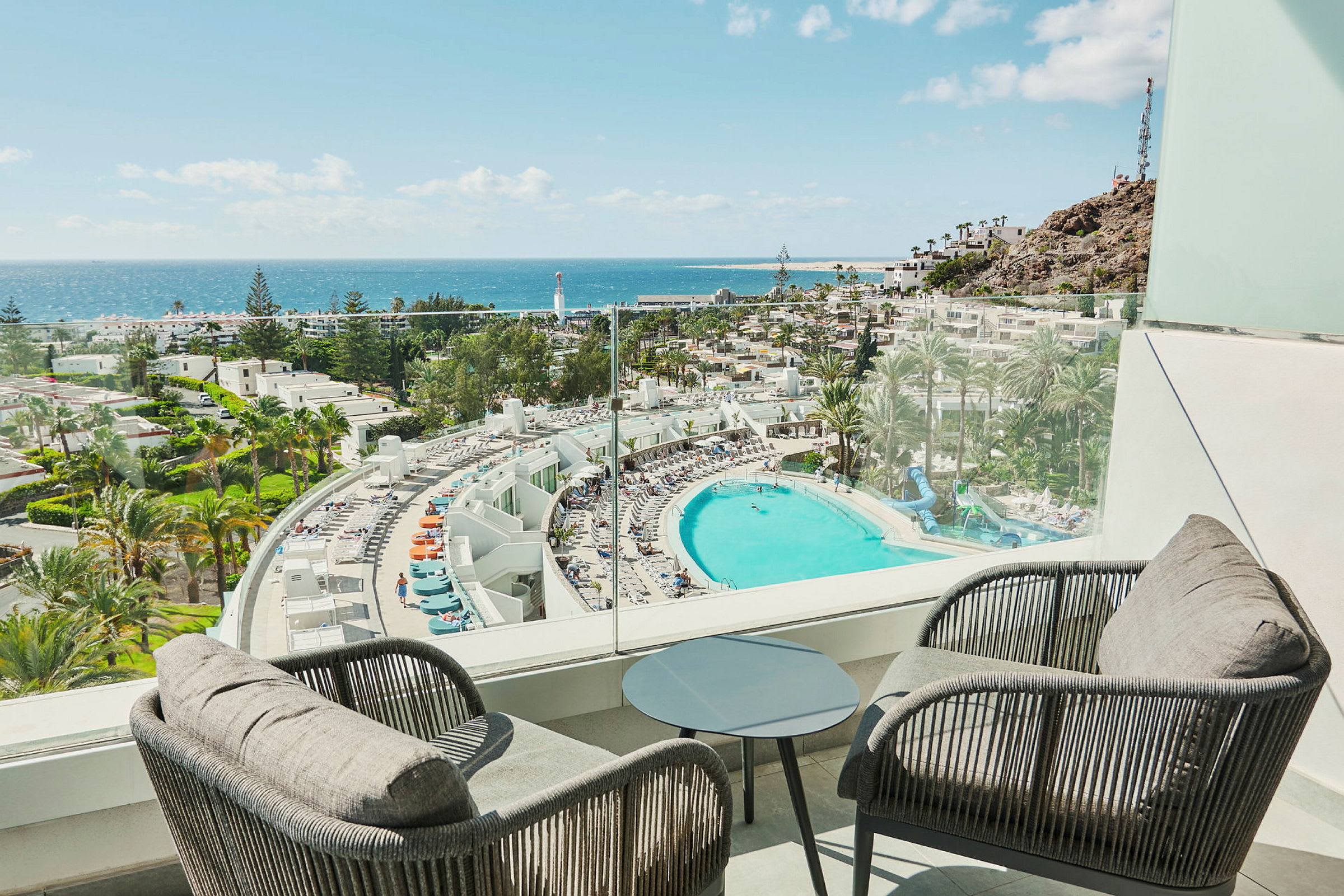 Utsikt från en balkong över ett hotell med en stor pool, omgivande palmer och havet i bakgrunden. (AI-genererad text)