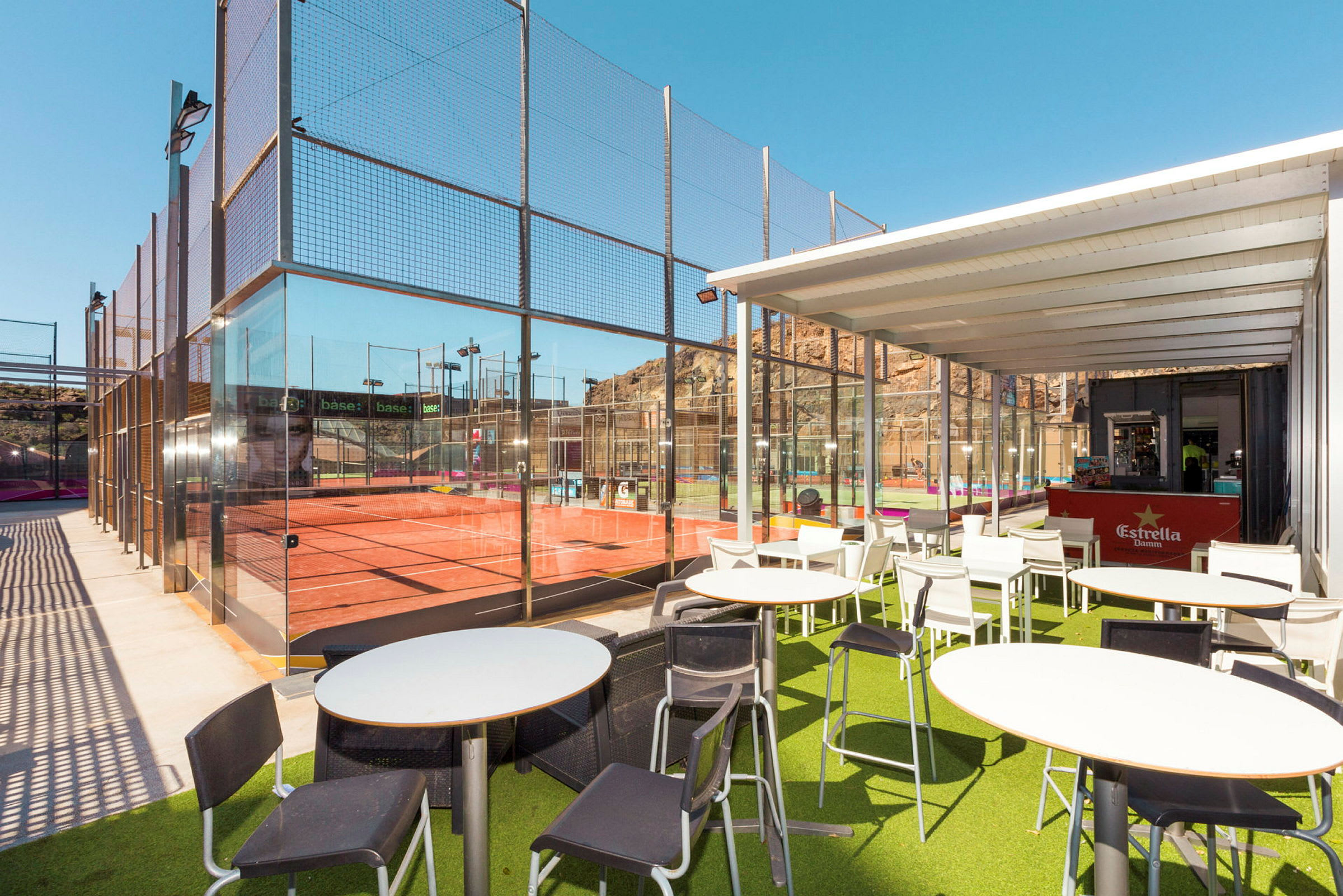 Uteservering vid en padelbana, med grönt konstgräs och vita möbler under ett tak. (AI-genererad text)