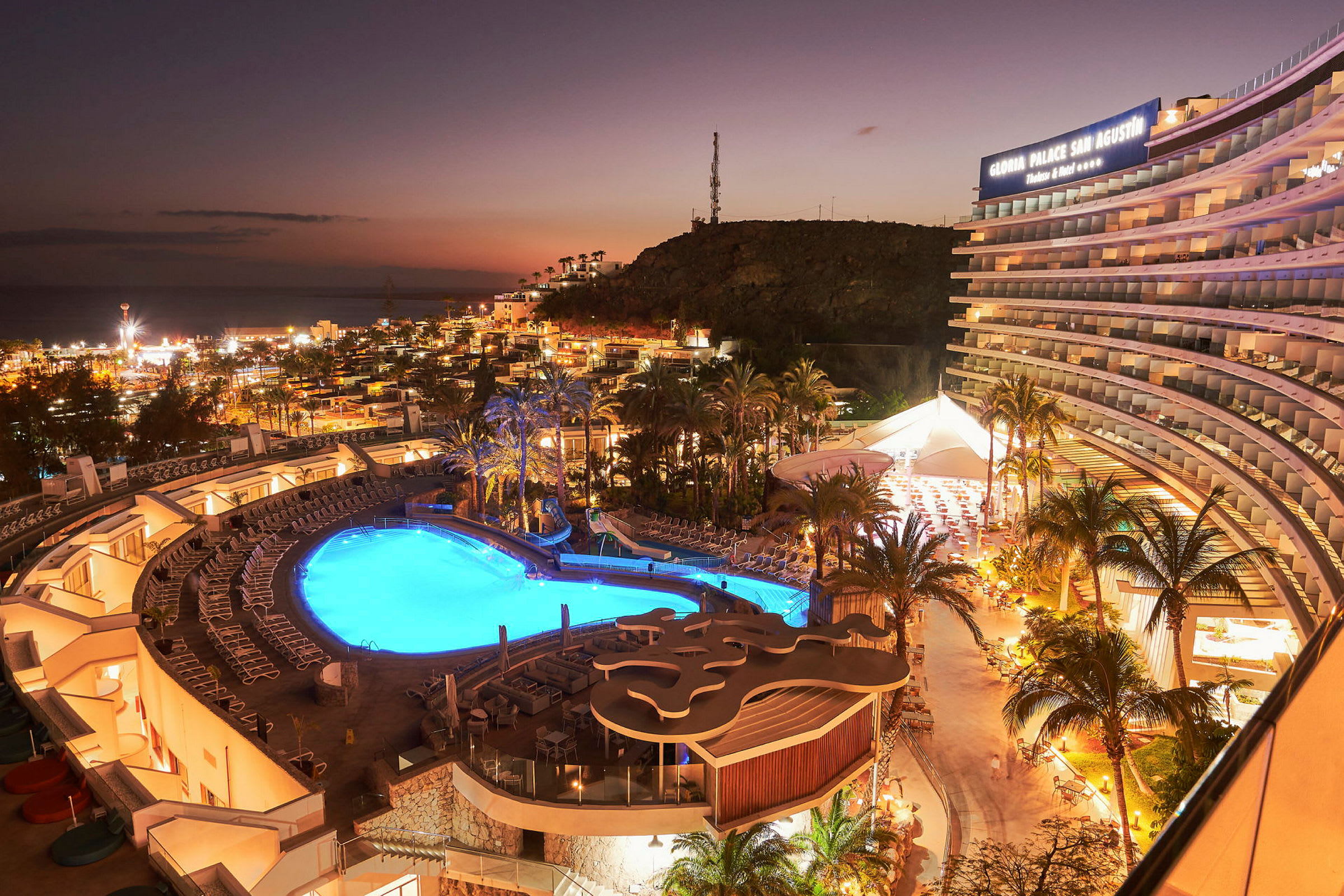 Hotellområde med upplyst pool och framträdande byggnad vid solnedgång. (AI-genererad text)