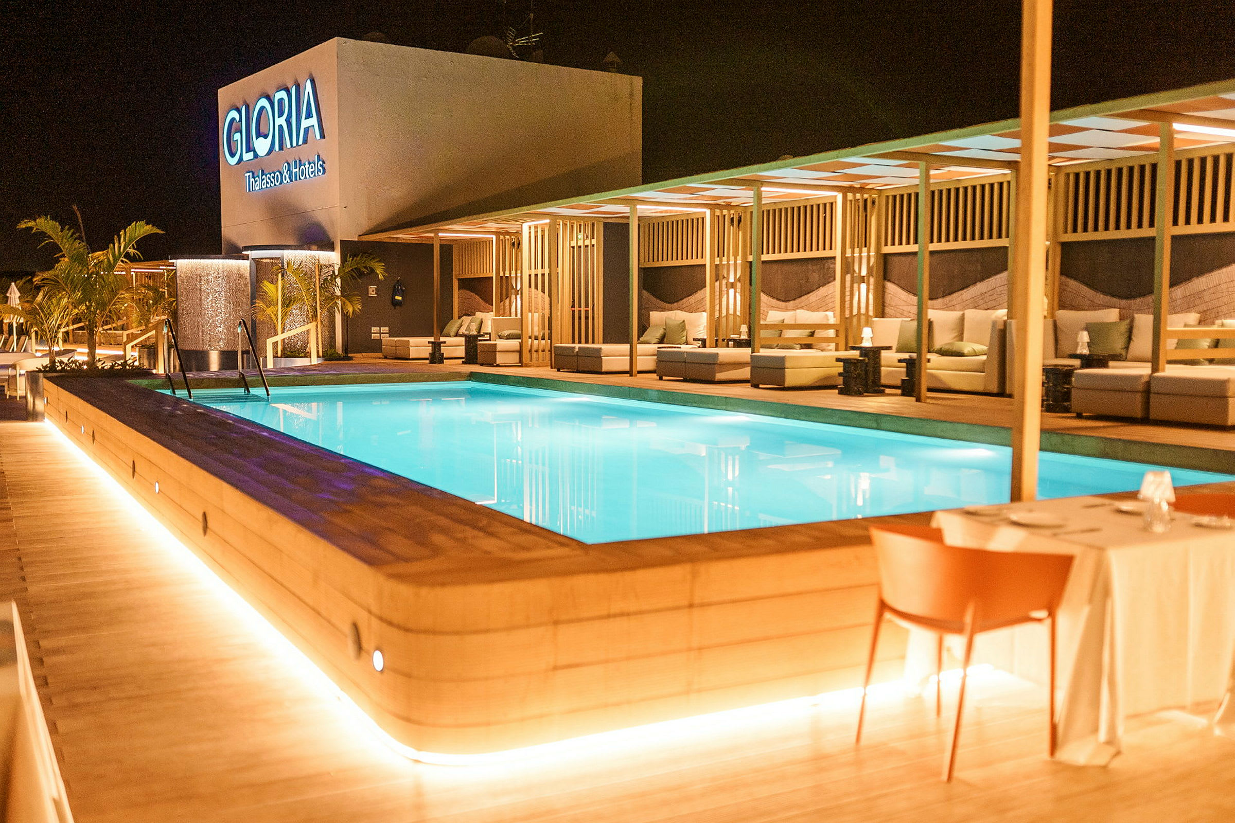 En upplyst swimmingpool omgiven av moderna sittplatser och bord på ett hotellområde. (AI-genererad text)