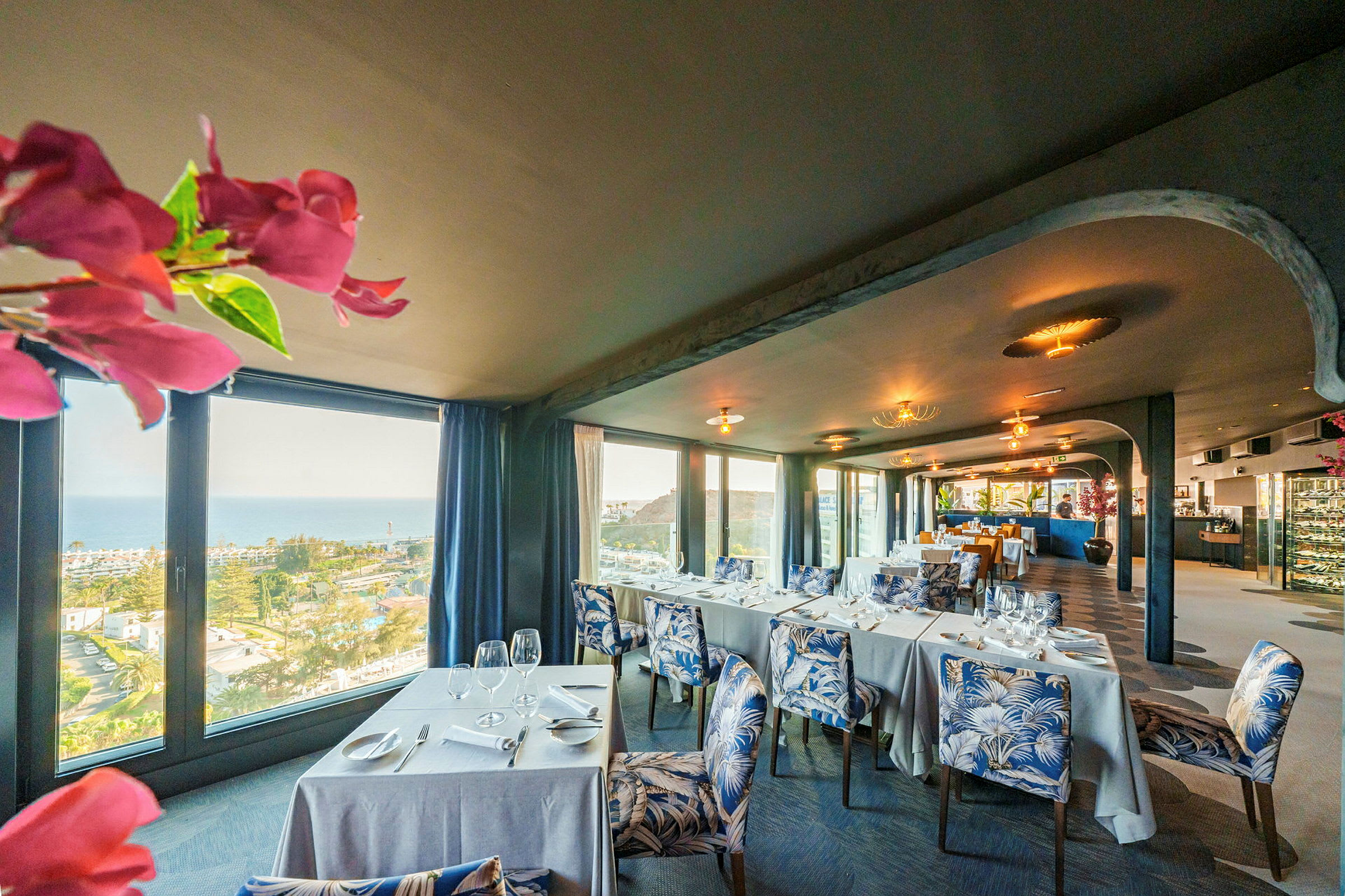 Utsikt över en elegant restaurang med bord klädda i vita dukar, stolar med bladmönster och stora fönster med utsikt över hav och landskap. (AI-genererad text)