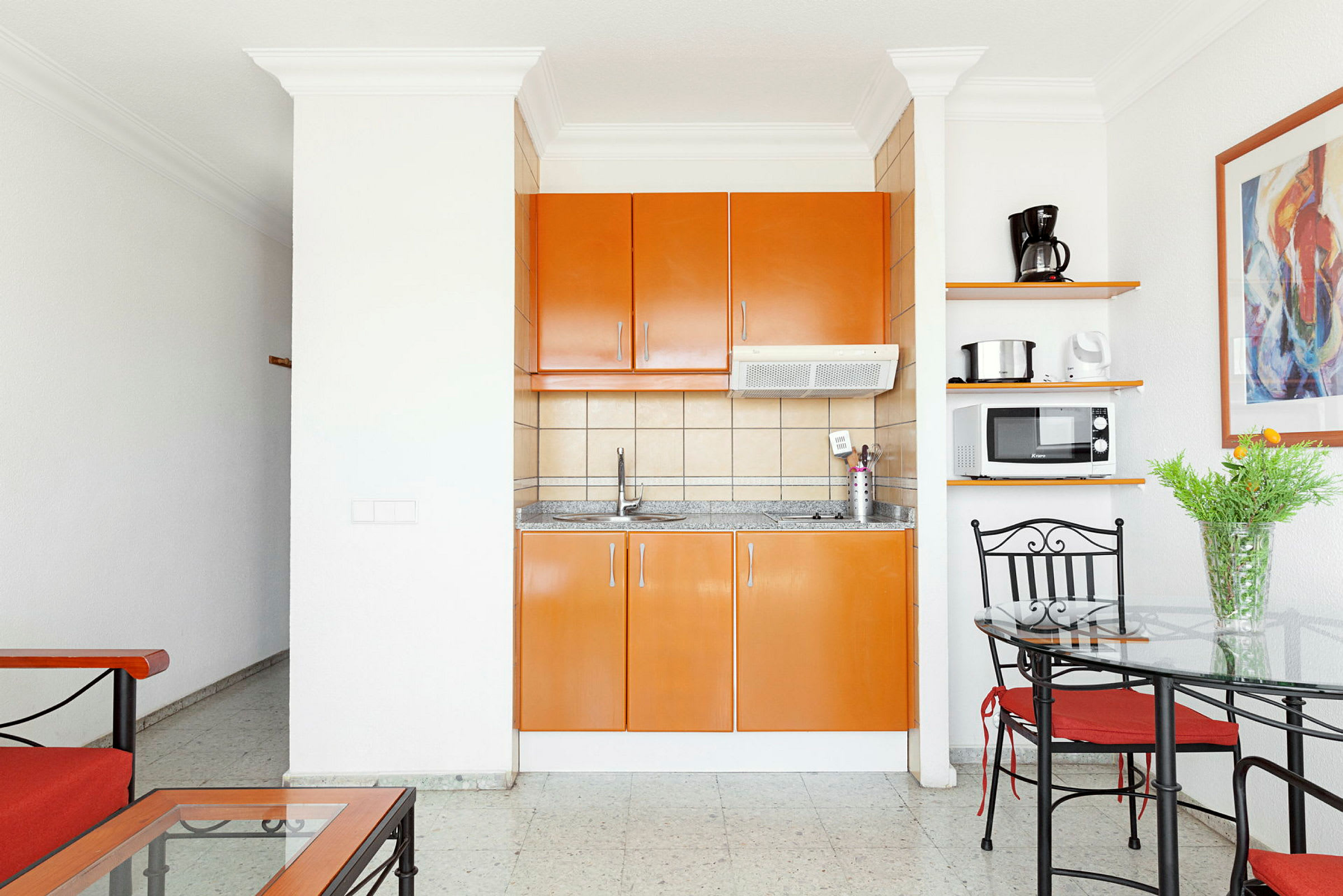Ett kök med orange skåp, en diskbänk och hushållsapparater, bredvid en matplats med glasbord och metallstolar. (AI-genererad text)