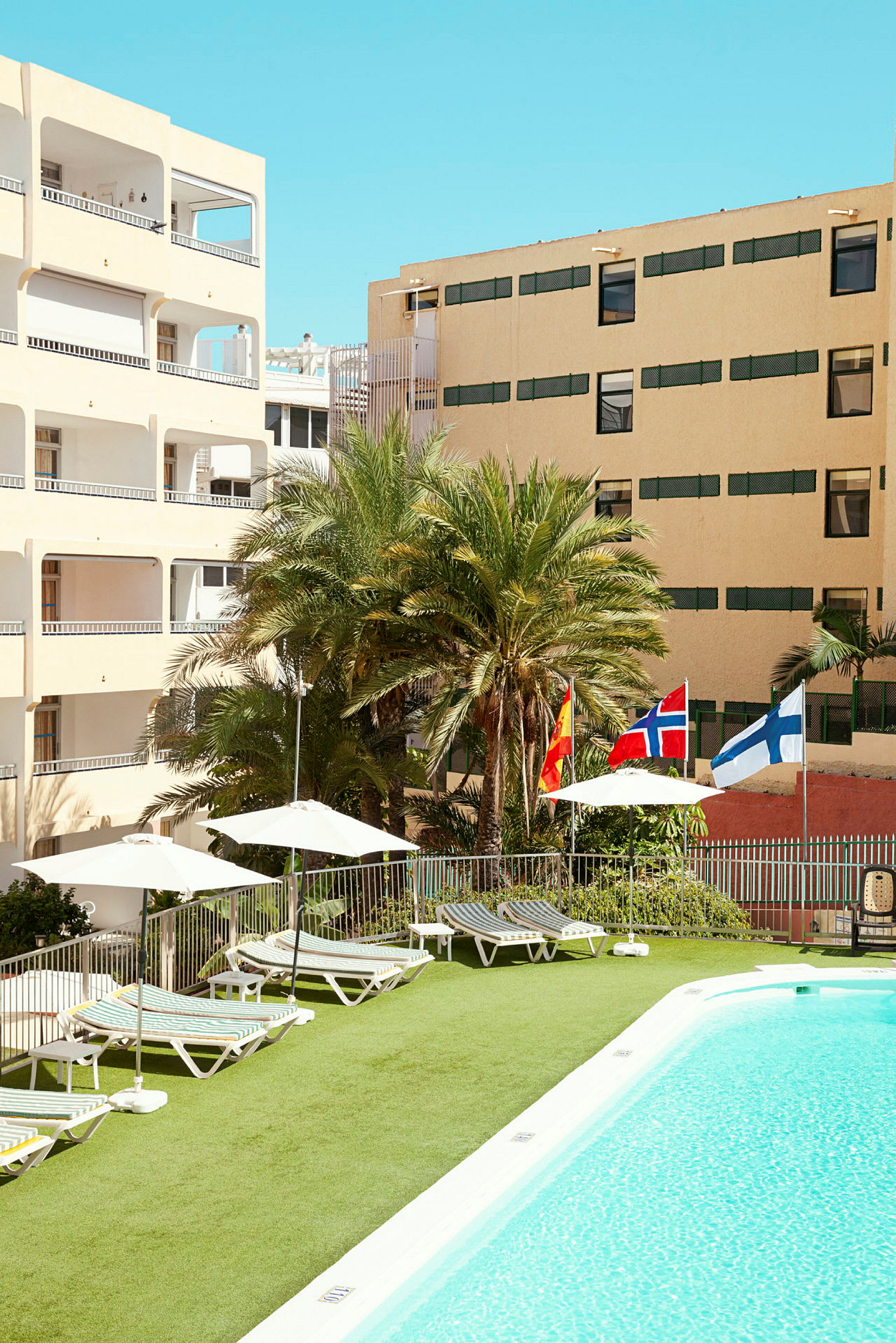 Bild av en hotellpool med solstolar, parasoller och flaggor från Spanien, Norge och Finland. Bakgrunden visar hotellbyggnader och palmer. (AI-genererad text)